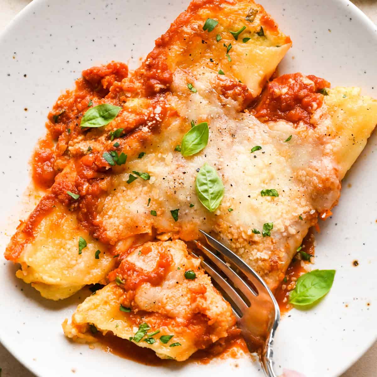 Close-up view of stuffed manicotti topped with fresh basil and cracked pepper, being served on a white speckled plate with a fork.
