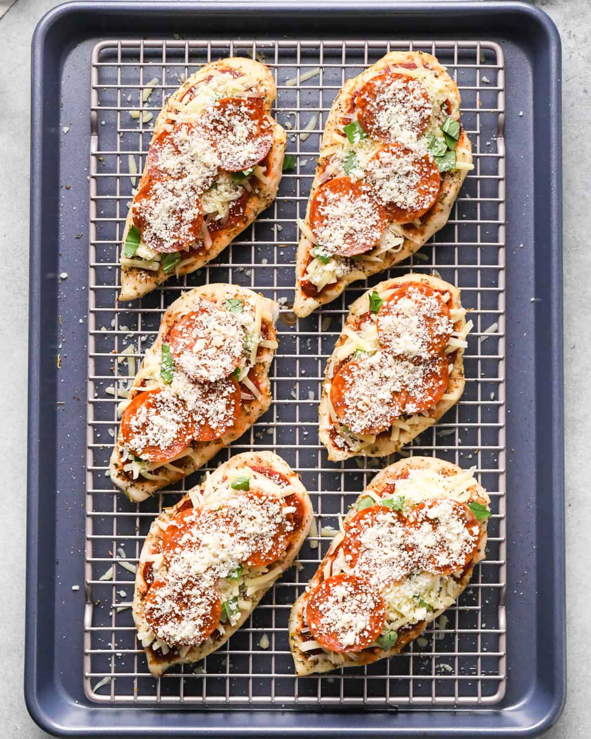 Making pizza chicken: overhead photo of chicken breasts topped with spices, pizza sauce, mozzarella cheese, Parmesan cheese, pepperoni, and fresh basil leaves.