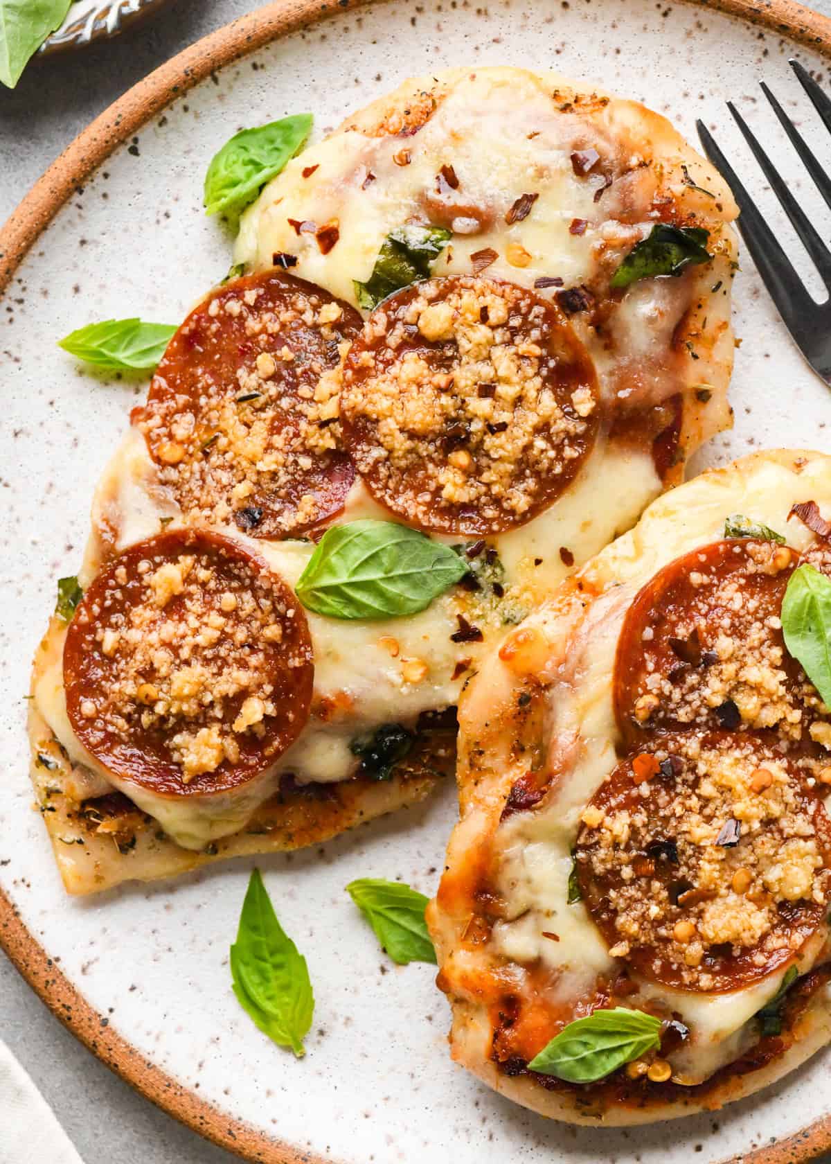 Plate of baked pizza chicken topped with melted mozzarella, pepperoni slices, Parmesan cheese, and fresh basil leaves, with a fork sitting on the plate.