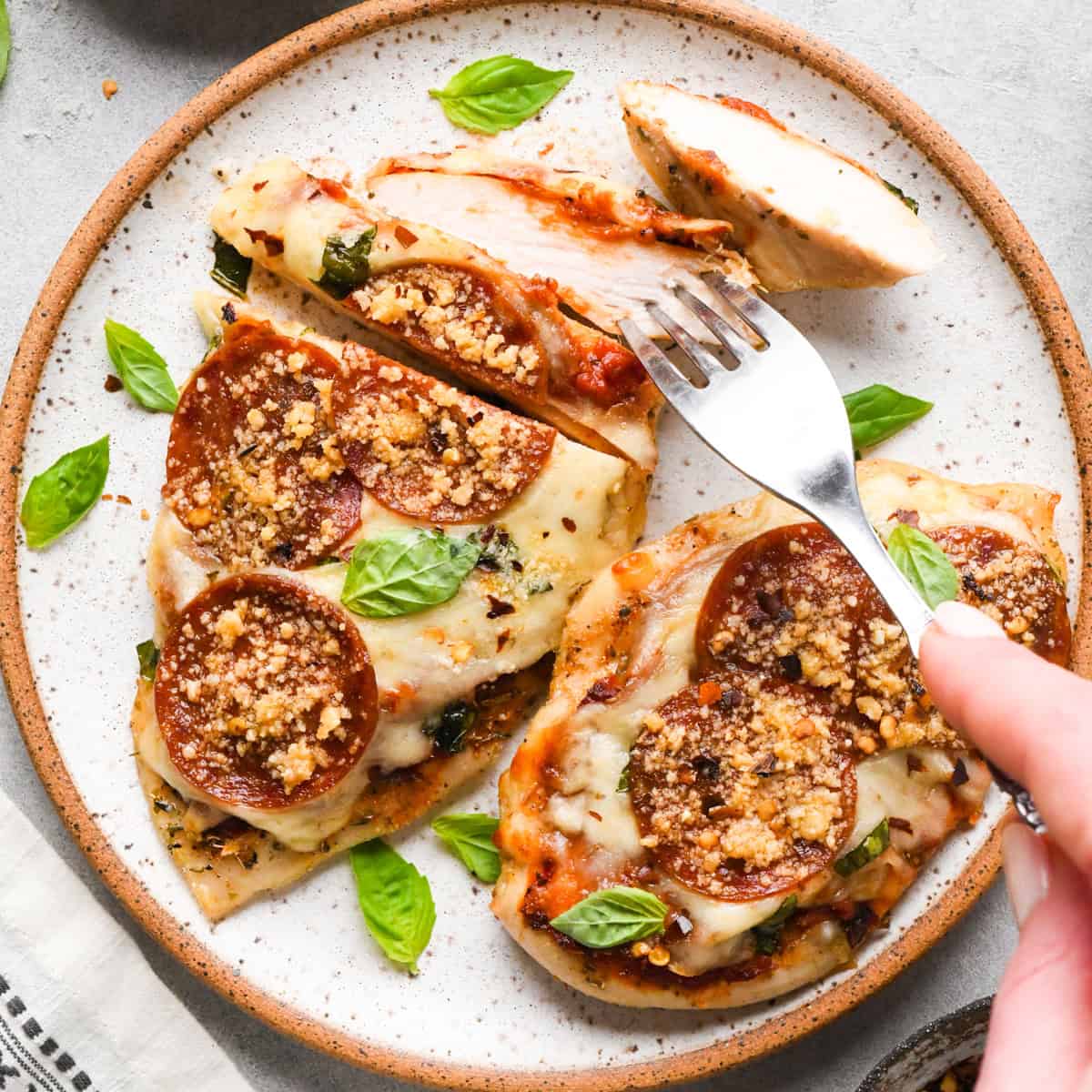 Plate of baked pizza chicken topped with melted mozzarella, pepperoni slices, Parmesan cheese, and fresh basil leaves, with a fork cutting into a piece.