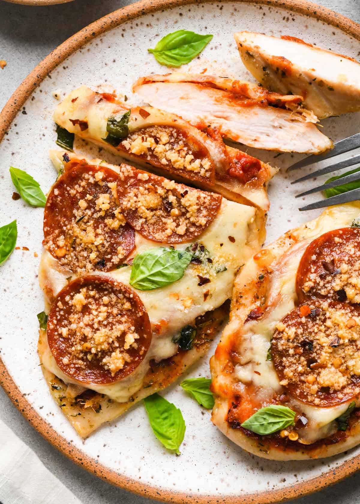 Plate of baked pizza chicken topped with melted mozzarella, pepperoni slices, Parmesan cheese, and fresh basil leaves, with a fork cutting into a piece.