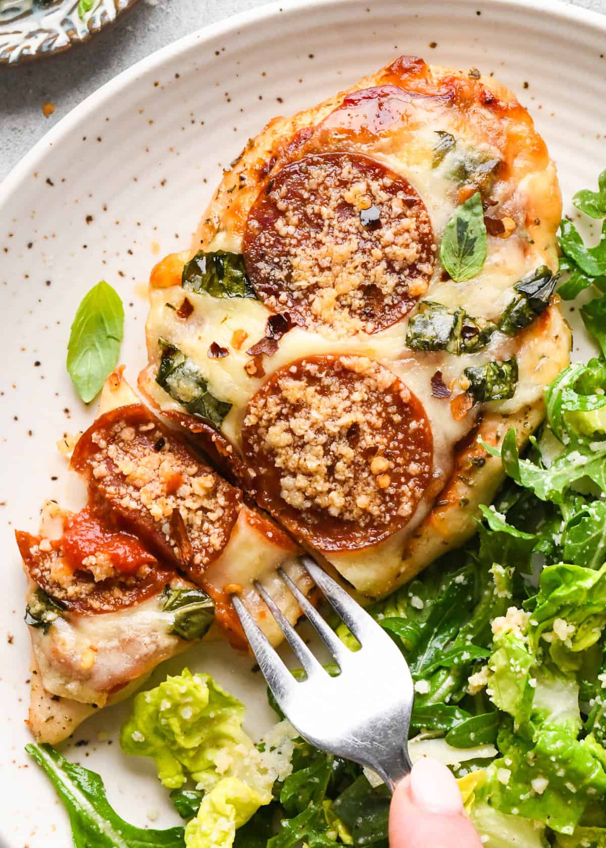 Close-up of baked pizza chicken topped with melted mozzarella, pepperoni slices, chopped basil, and grated Parmesan, served with a side of fresh green salad with a fork.