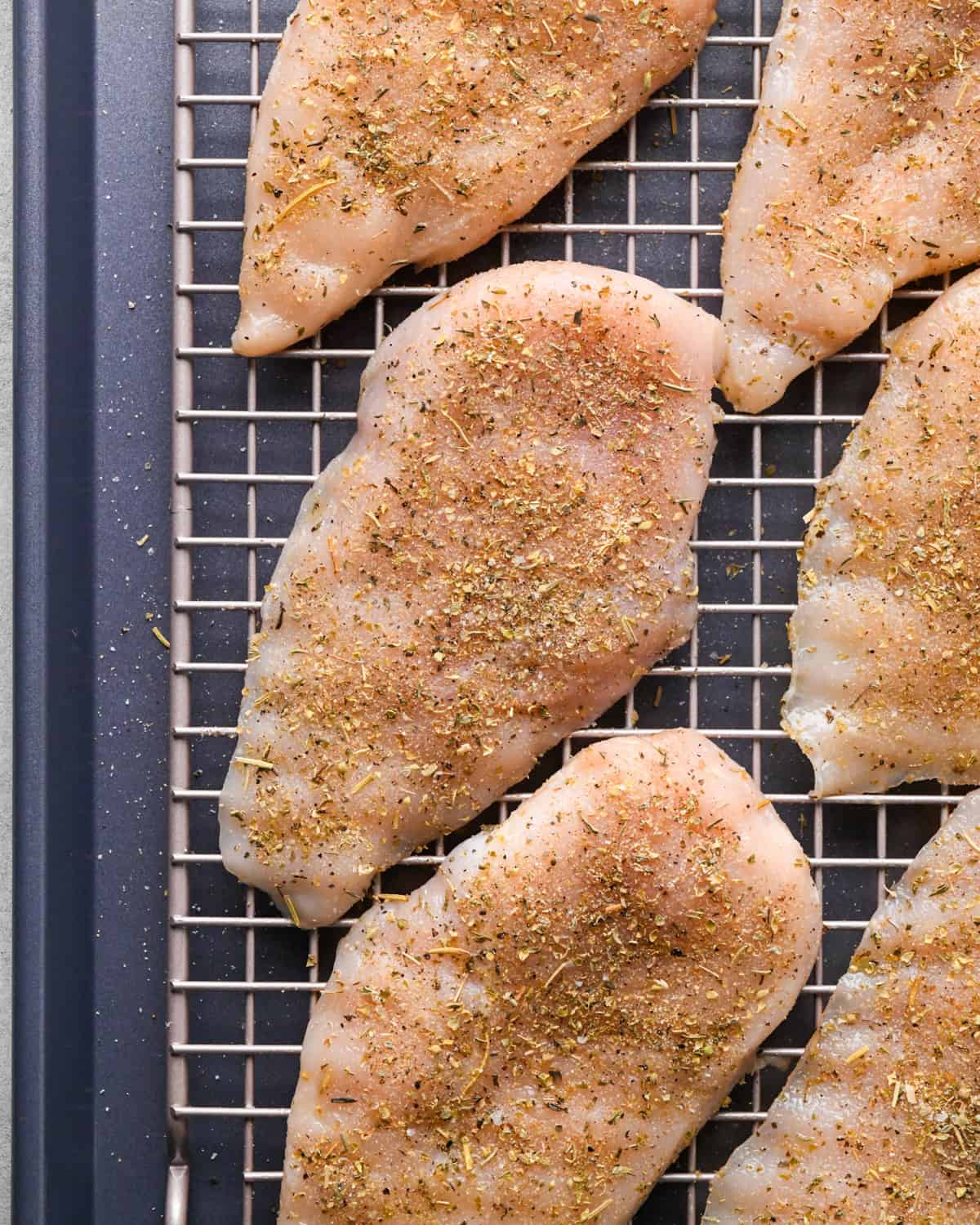 Making pizza chicken: close-up photo of chicken breasts sprinkled with spices and placed on a cooling rack on top of a baking sheet.