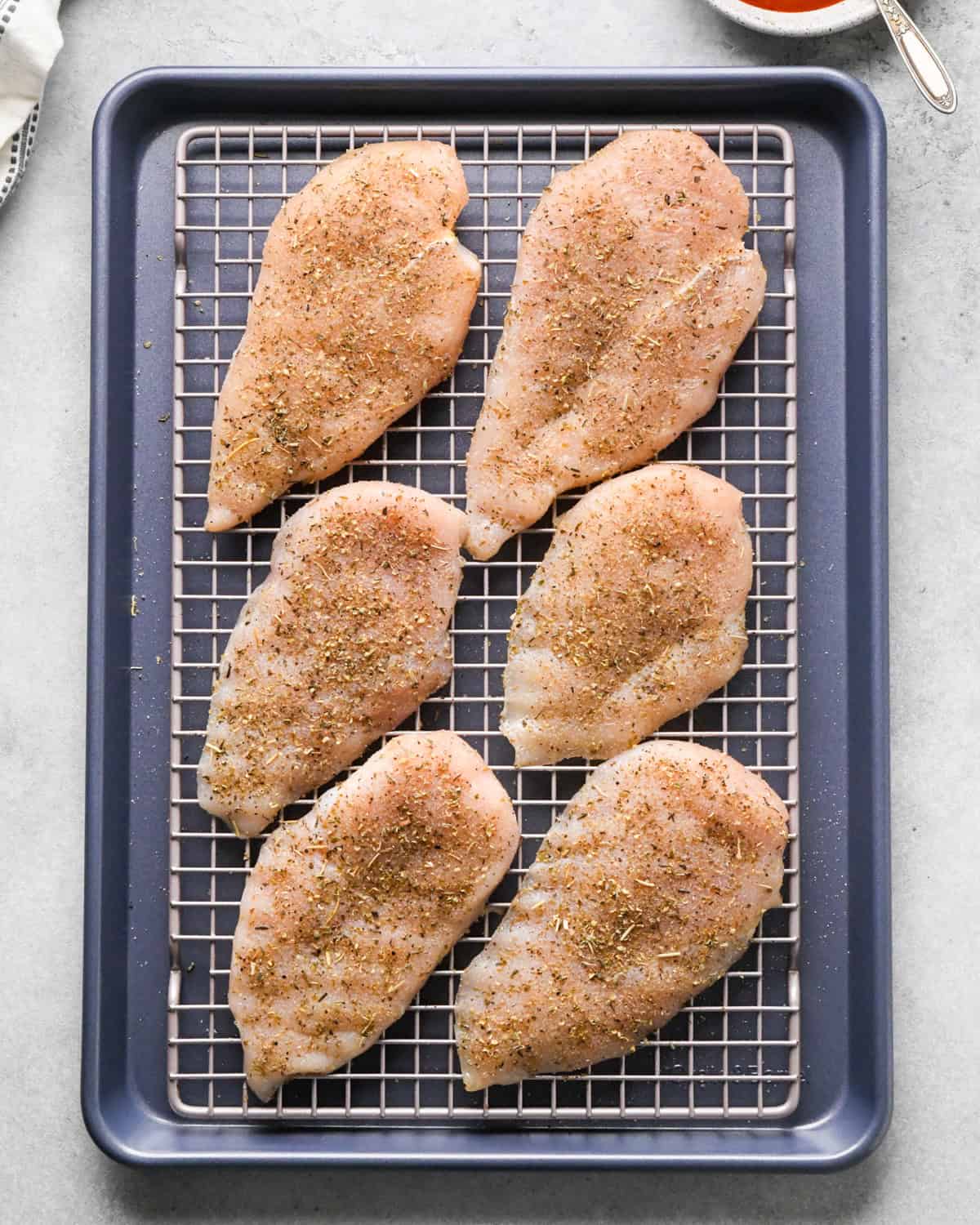 Making pizza chicken: overhead photo of chicken breasts sprinkled with spices and placed on a cooling rack on top of a baking sheet.