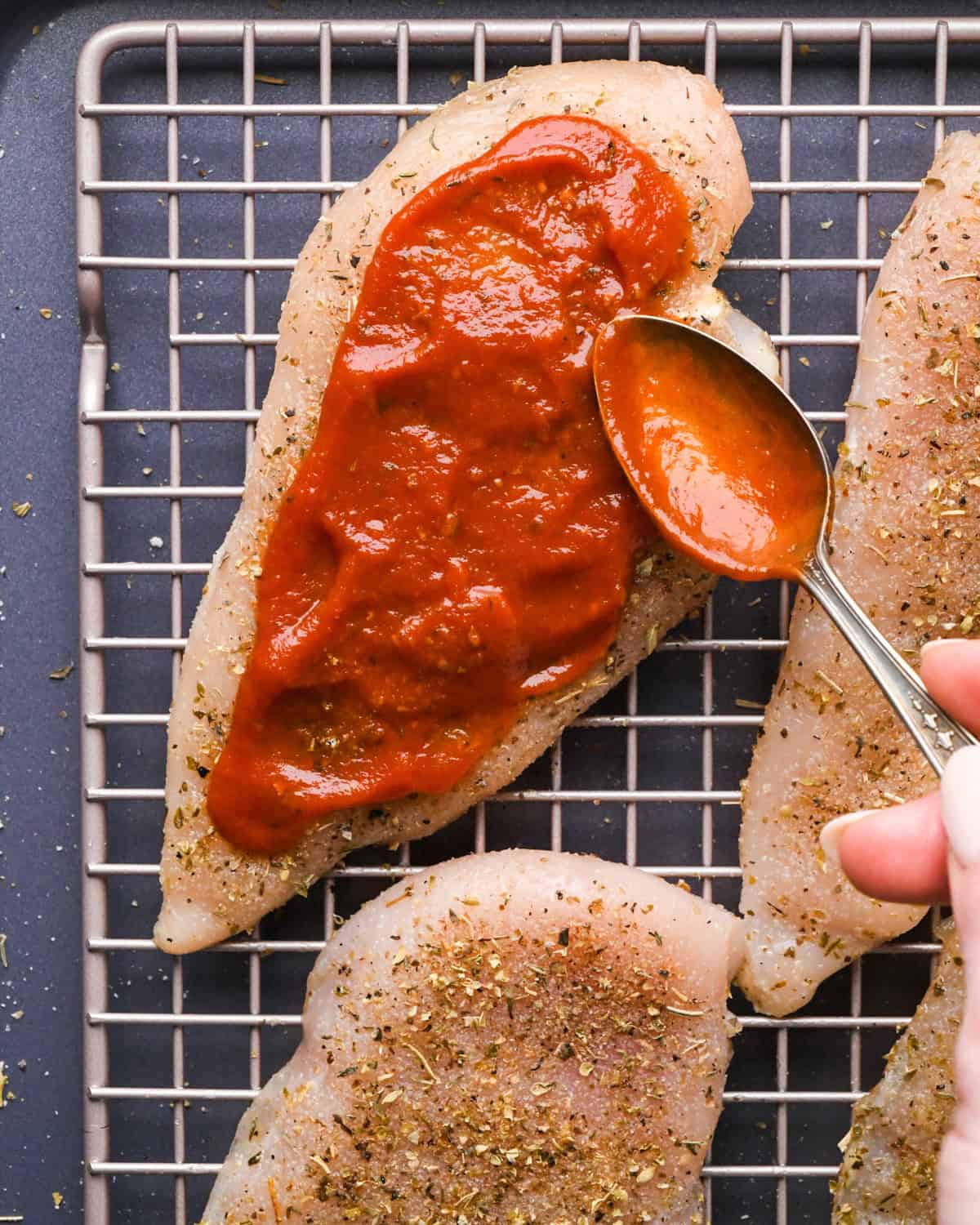 Making pizza chicken: close-up photo of chicken breasts sprinkled with spices and pizza sauce being spread on top.