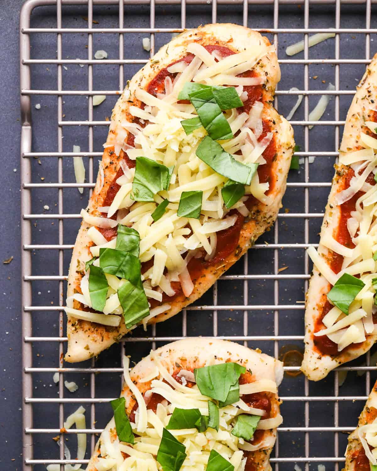 Making pizza chicken: overhead photo of chicken breasts topped with spices, pizza sauce, mozzarella cheese, and fresh basil leaves.