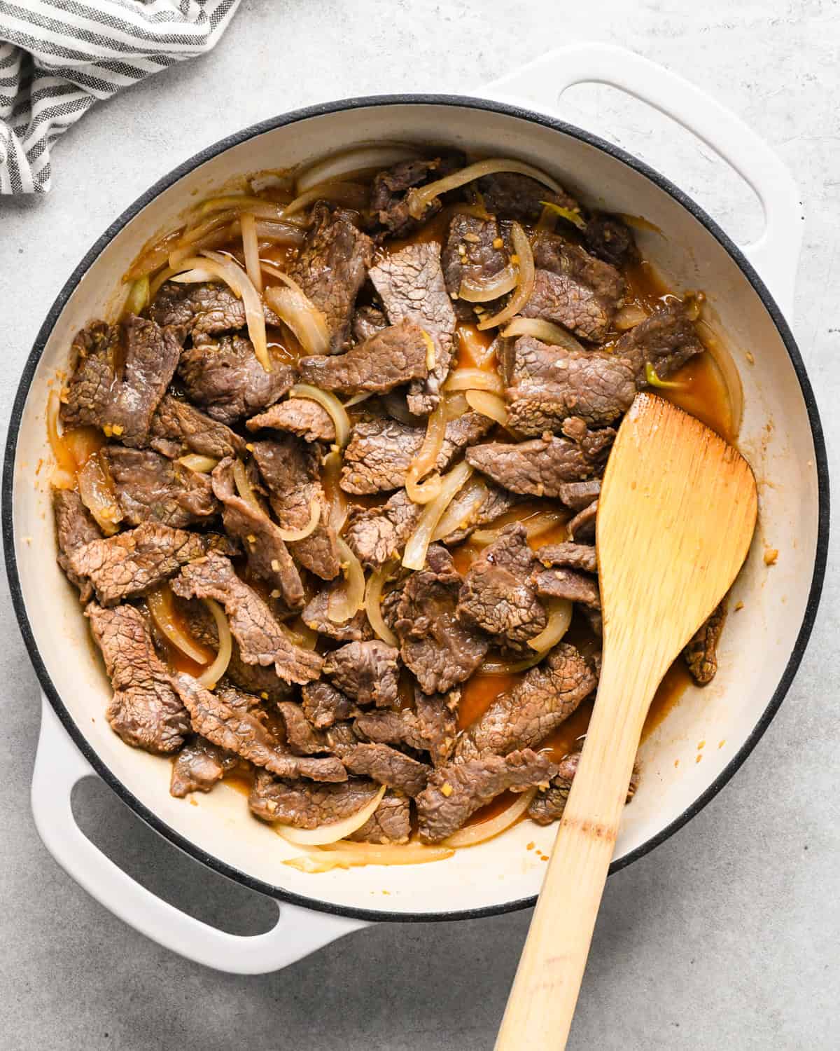 Making beef and broccoli: flank steak, onions, and spices are added to a white pan and sautéed with a wooden spatula.