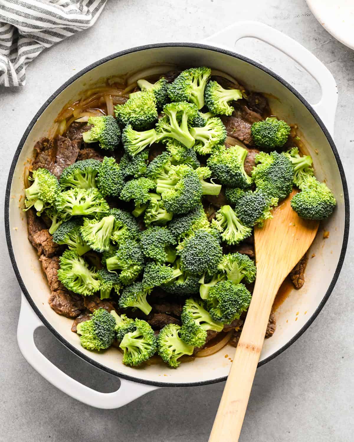 Making beef and broccoli: adding broccoli to a white pan with sautéed flank steak, onion, and spices. Food is being stirred with a wooden spatula.