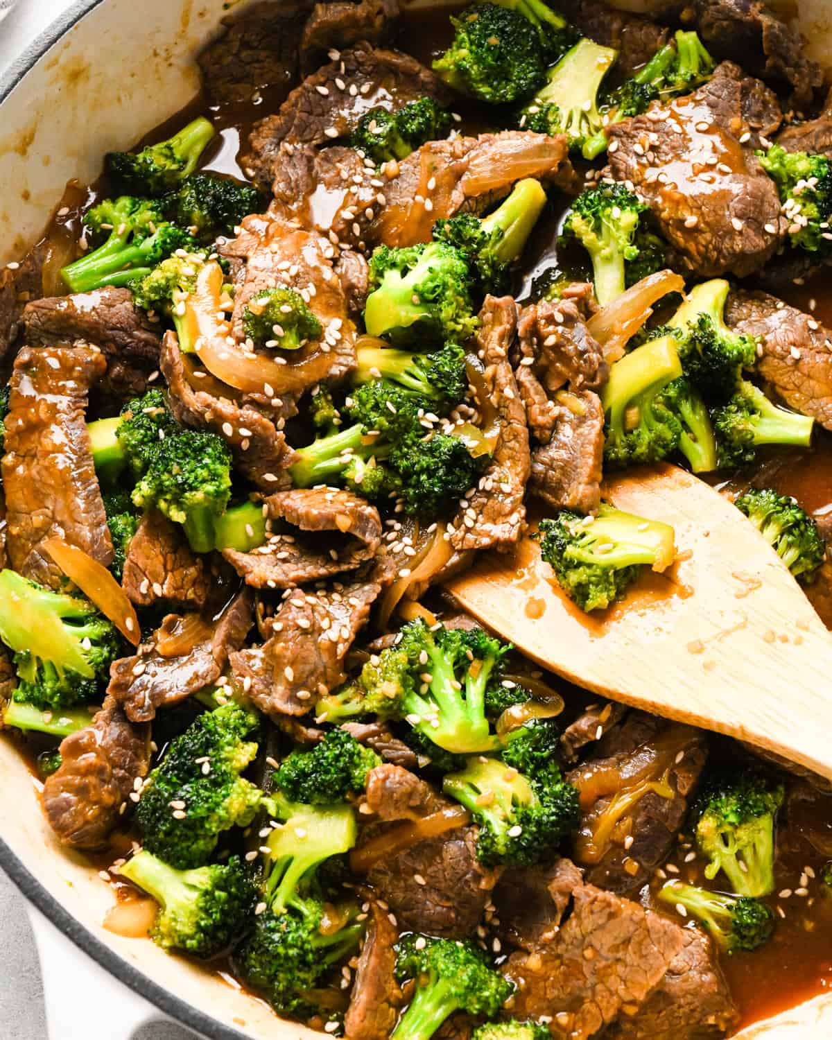 Overview photo of beef and broccoli stir-fry served over white rice with sesame seeds on top, being stirred with a wooden spatula.