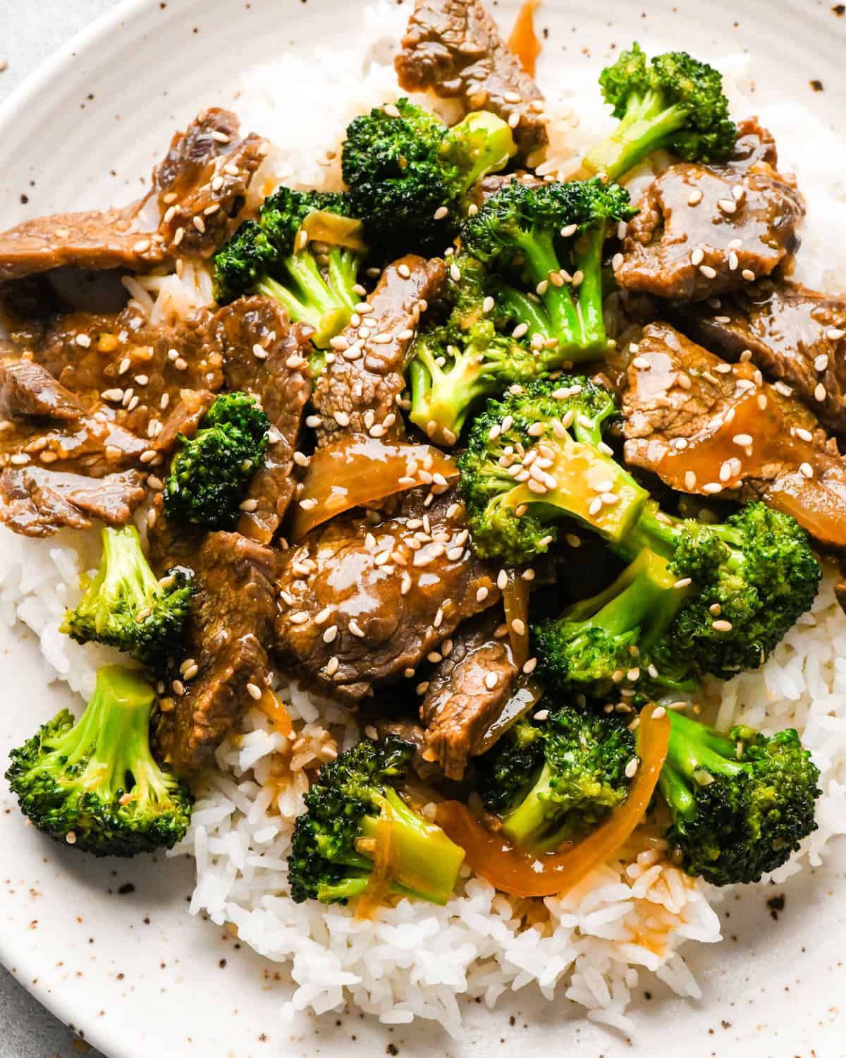 Making beef and broccoli: beef and broccoli stir-fry on top of white rice and sprinkled with sesame seeds, being served on a white speckled plate.