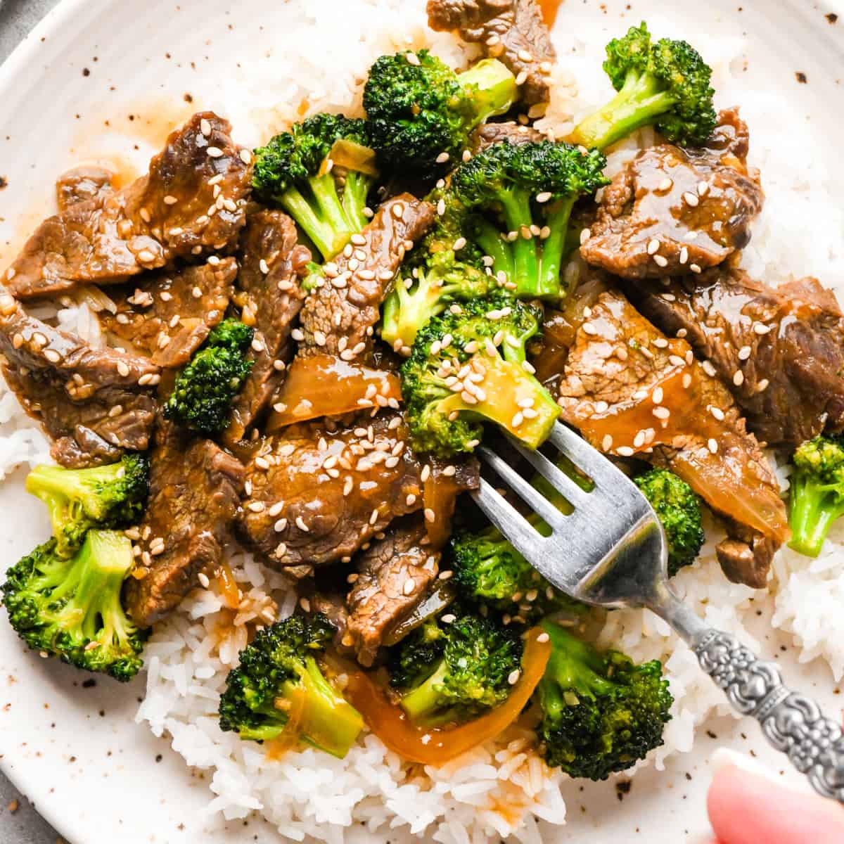 Making beef and broccoli: beef and broccoli stir-fry on top of white rice and sprinkled with sesame seeds, being served on a white speckled plate with a fork.