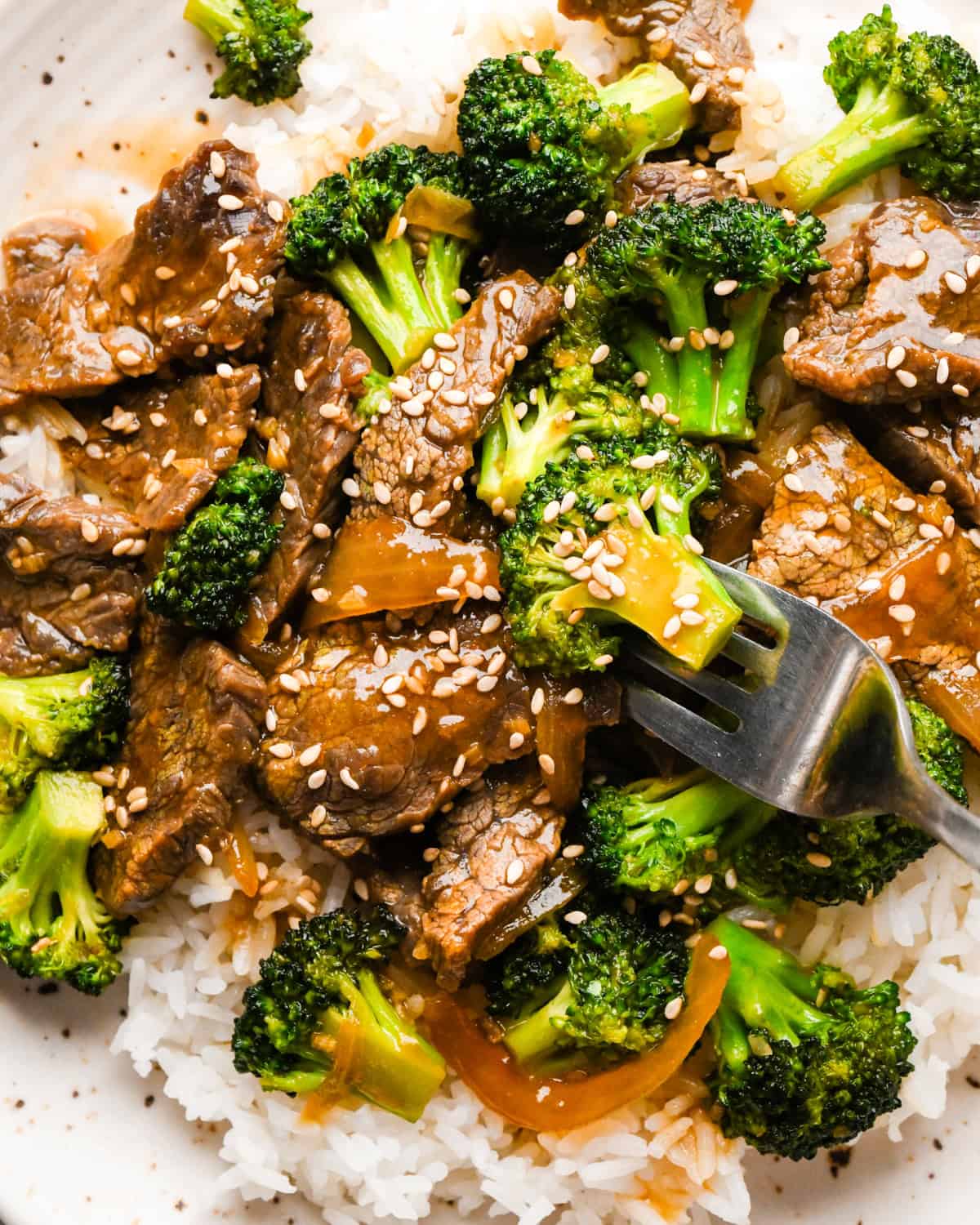 Overview photo of beef and broccoli stir-fry served over white rice with sesame seeds on top, being eaten with a fork.