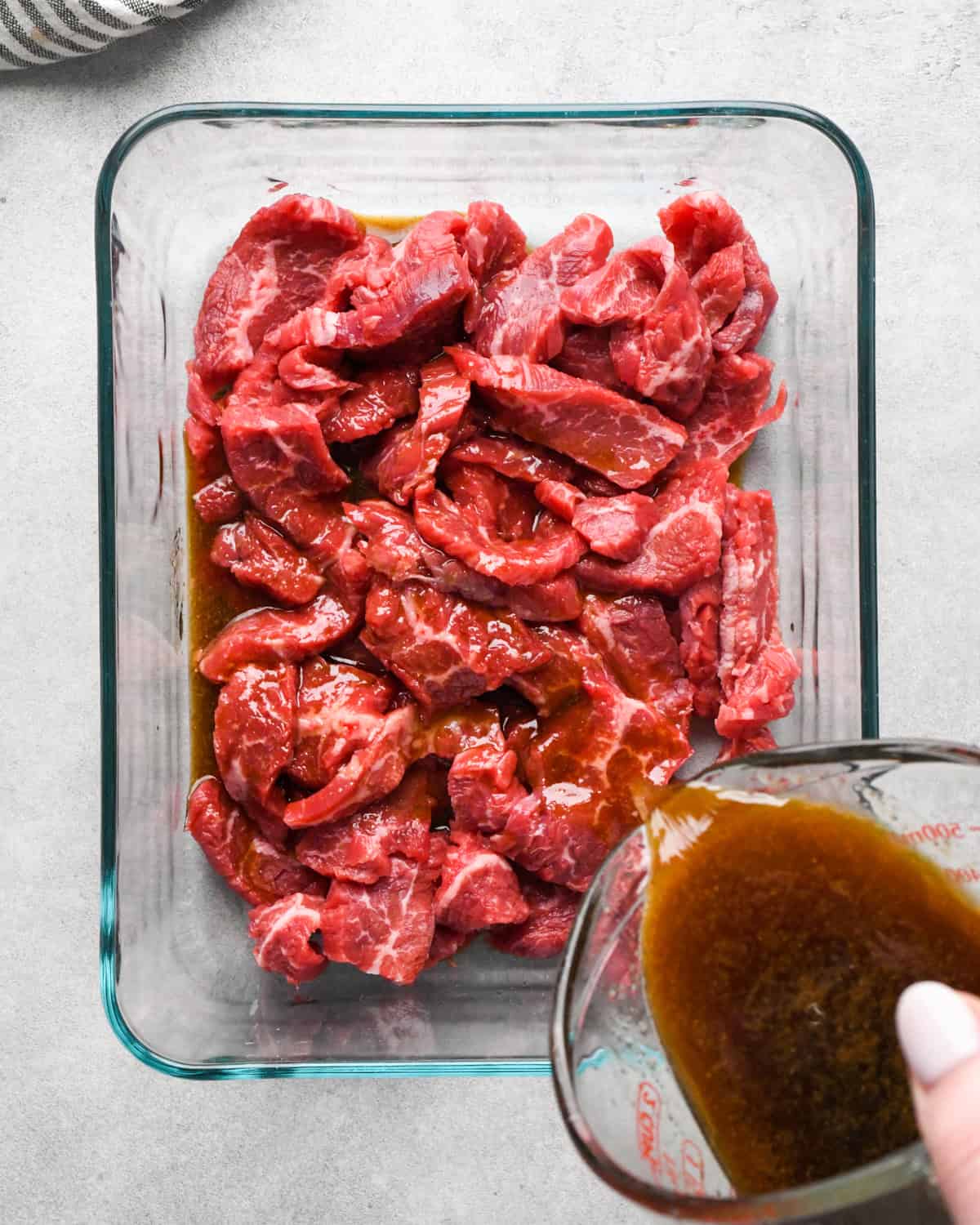 Making beef and broccoli: overview photo of sauce being poured over flank steak in a clear rectangle serving dish.