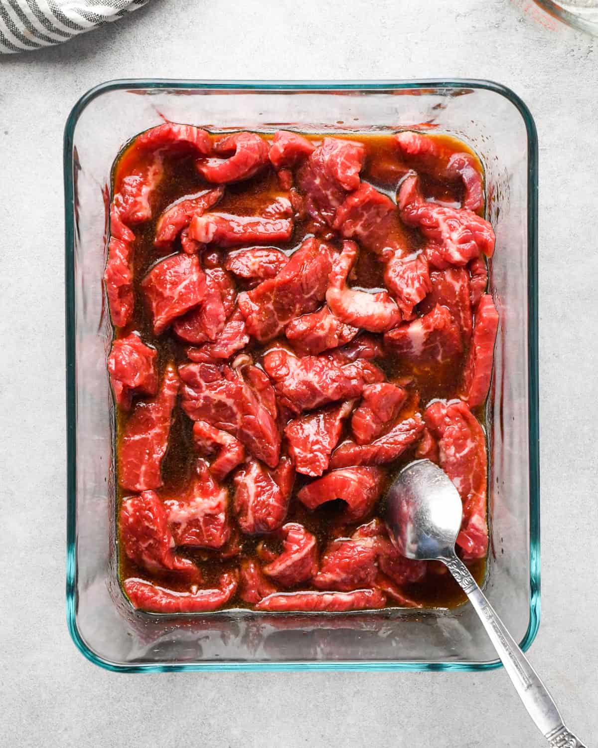 Making beef and broccoli: overview photo of sauce being poured over flank steak in a clear rectangle serving dish and mixed together with a spoon.