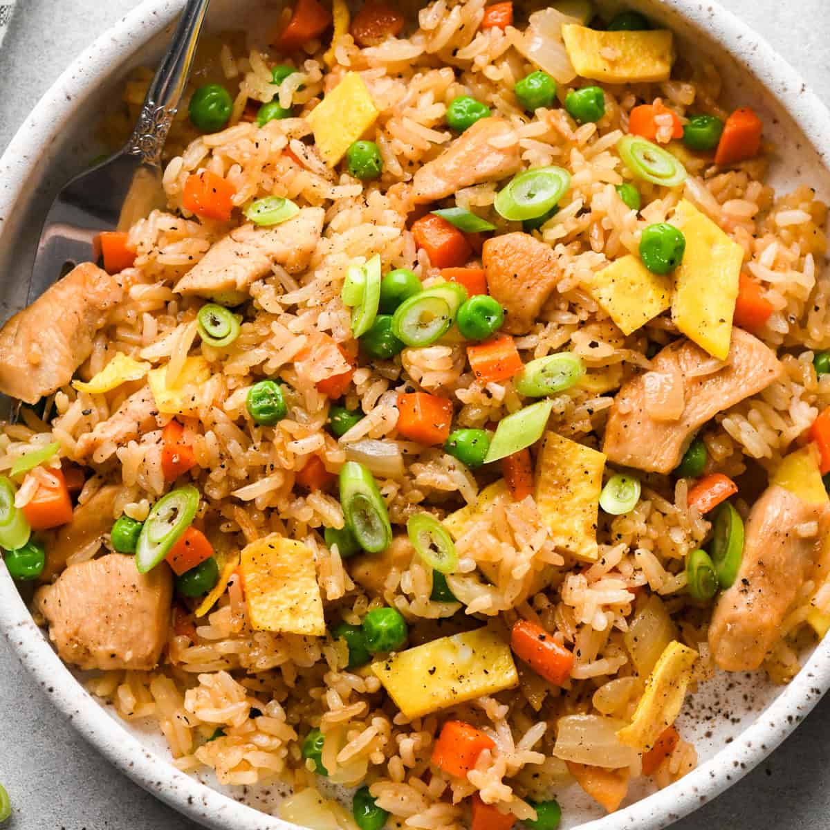 Bowl of chicken fried rice with diced chicken, peas, carrots, scrambled egg pieces, and sliced green onions, seasoned with soy sauce and black pepper and served in a white speckled bowl.