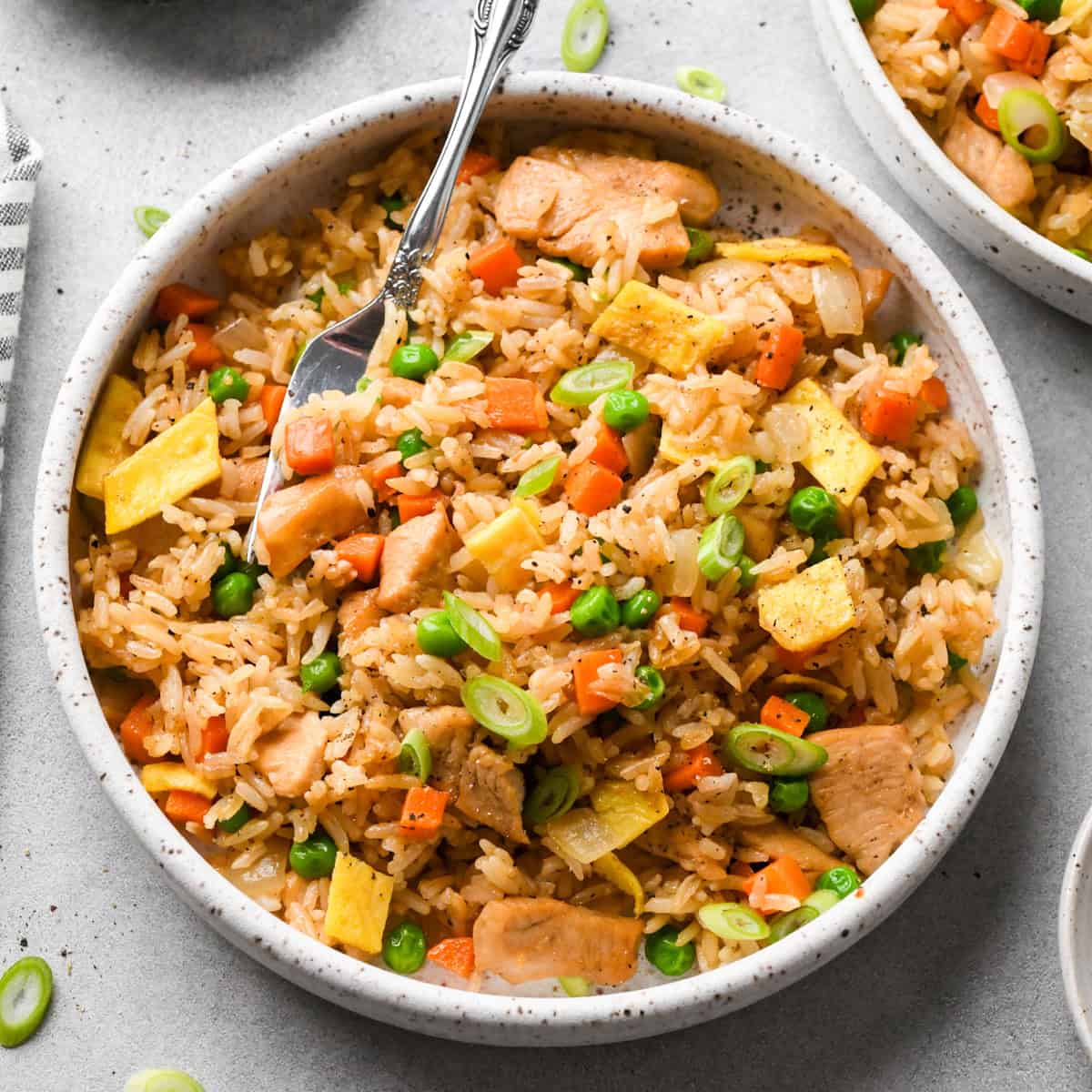 A bowl of chicken fried rice filled with pieces of chicken, scrambled egg, peas, carrots, onions, and green onions, with a fork resting inside the bowl on a light gray surface.