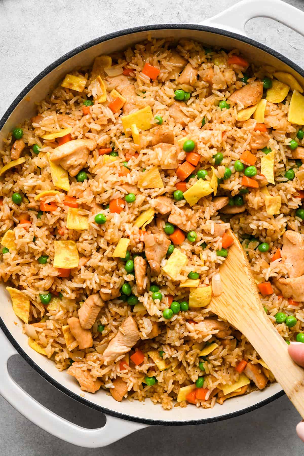 A skillet of chicken fried rice with diced chicken, peas, carrots, scrambled egg pieces, and sliced green onions, seasoned with soy sauce and black pepper being served with a wooden spatula.