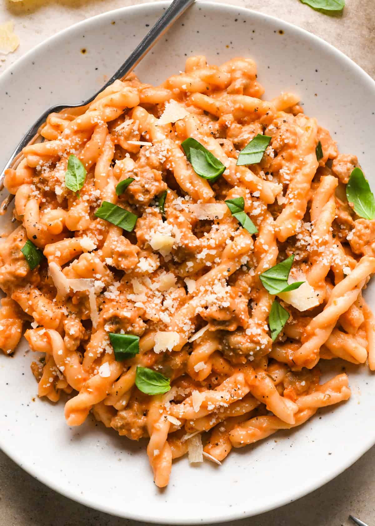 A white speckled bowl of creamy sausage pasta topped with grated Parmesan cheese and fresh basil leaves, being served with a fork.