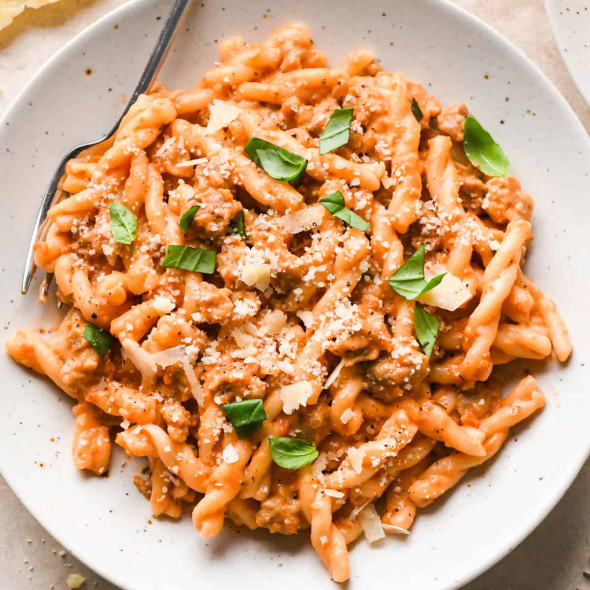 A plate of creamy sausage pasta topped with grated Parmesan cheese and fresh basil leaves, served with a fork on the side.