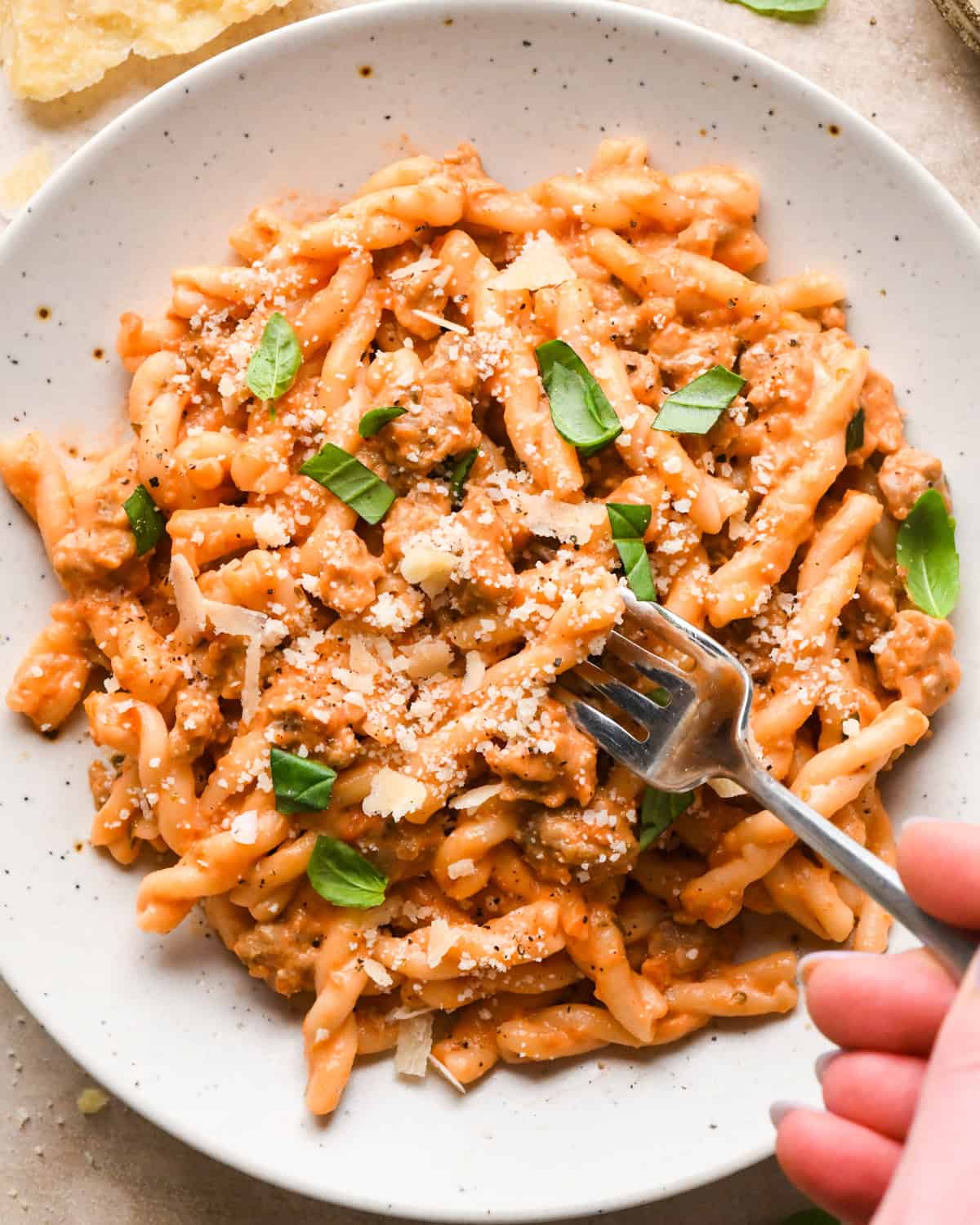 A white speckled bowl of creamy sausage pasta topped with grated Parmesan cheese and fresh basil leaves, being served with a fork.