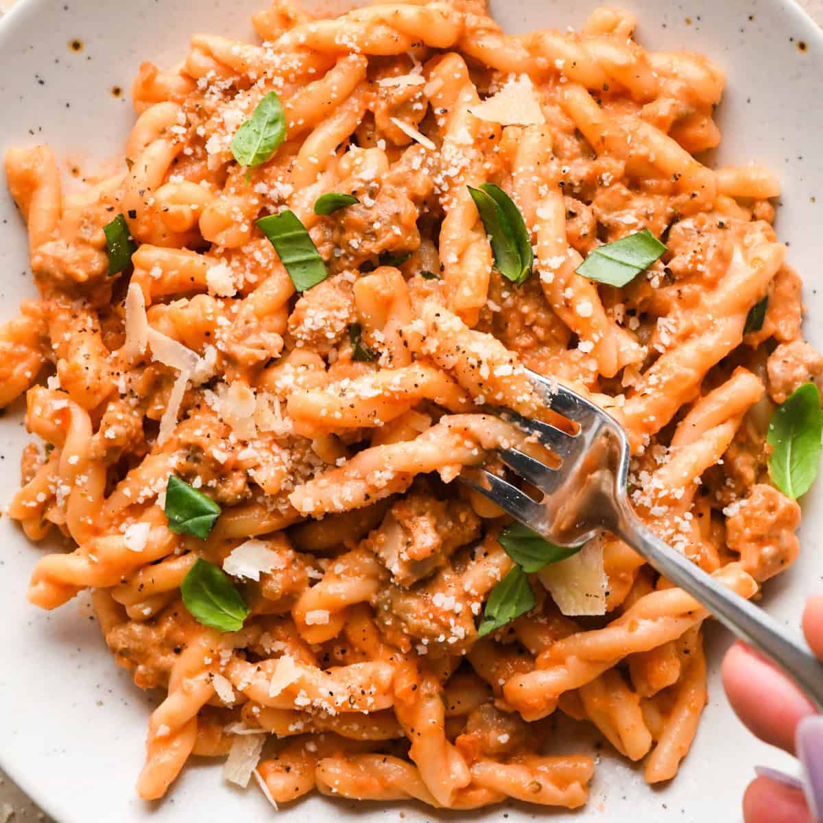 A plate of creamy sausage pasta topped with grated Parmesan cheese and fresh basil leaves, served with a fork on the side.