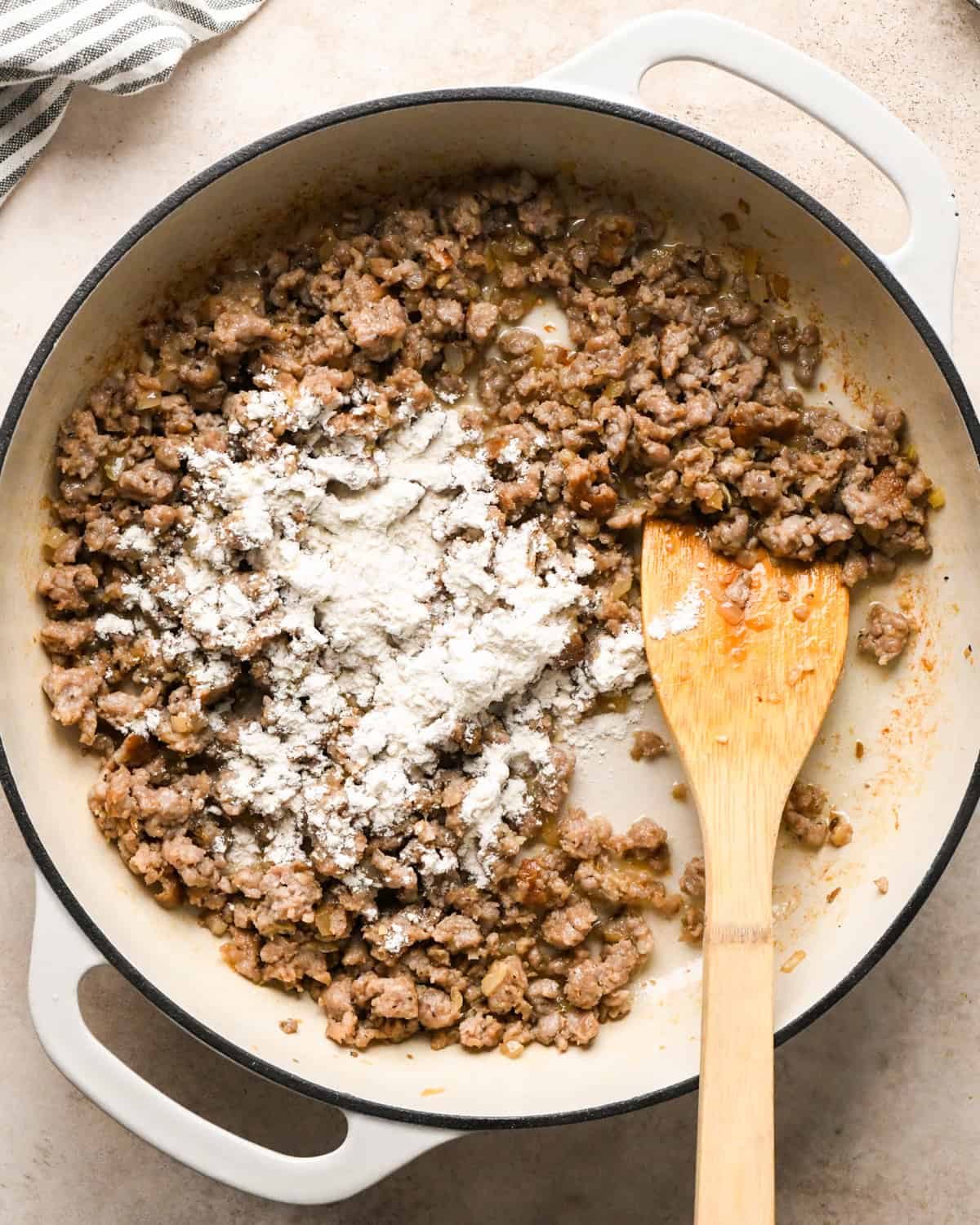 Making sausage pasta: overhead photo of Italian sausage, garlic, and onion cooked in a white skillet and being stirred with a wooden spatula. Flour has just been added.