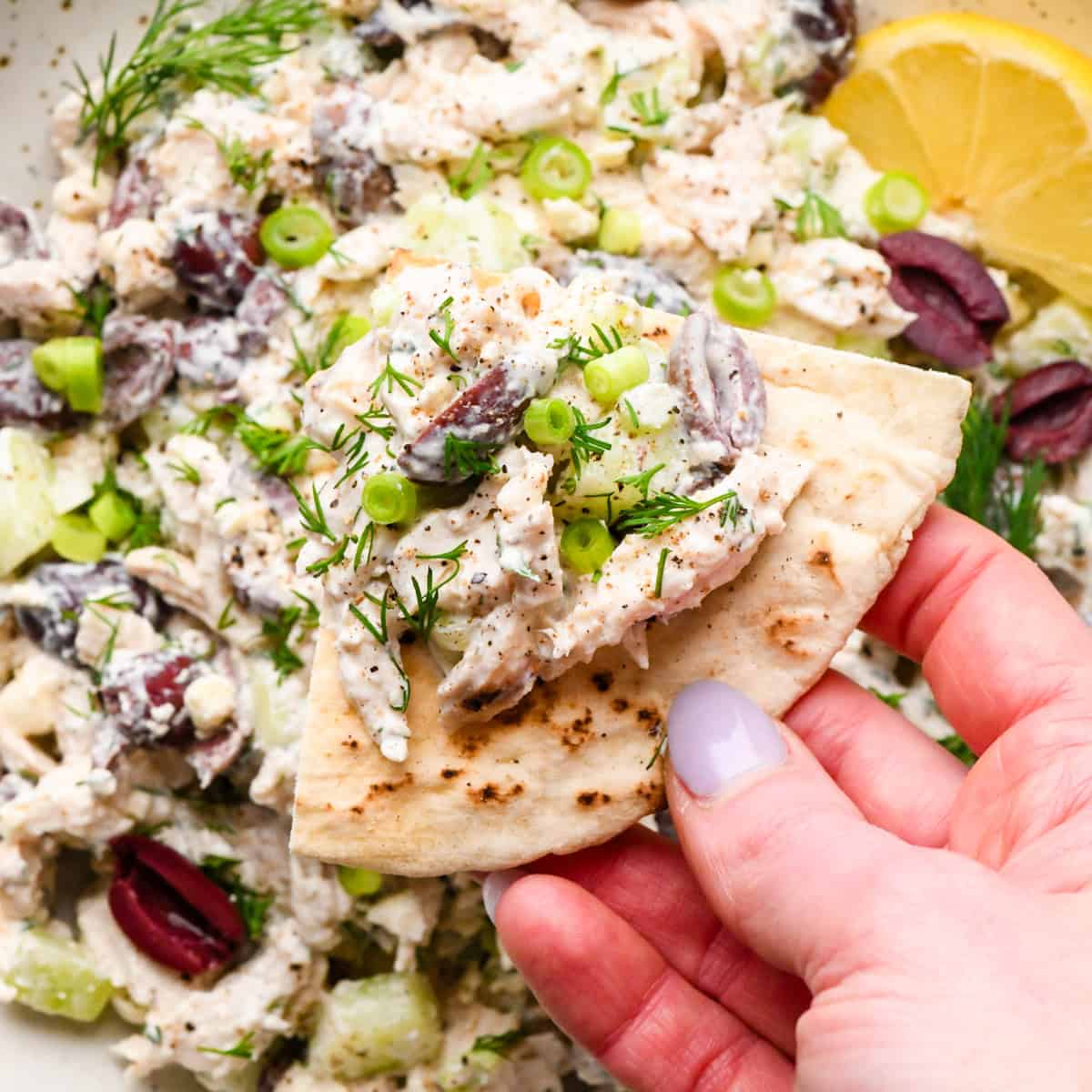 Overview photo of Greek chicken salad being eaten in a white speckled bowl with a slice of pita bread. A person's hand is holding the pita bread with some of the chicken salad on top.