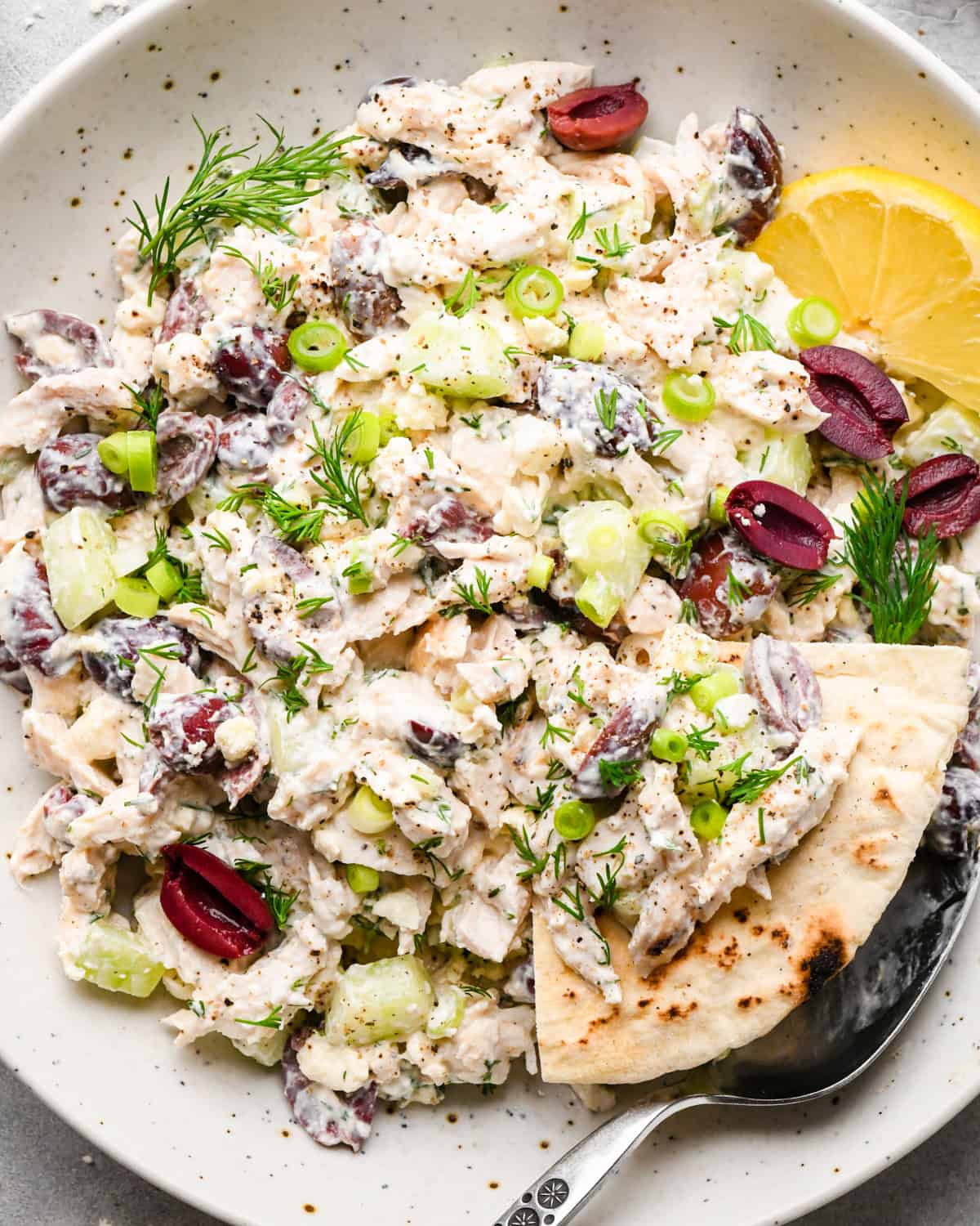 Overview photo of Greek chicken salad in a white speckled bowl with a slice of pita bread and a spoon.