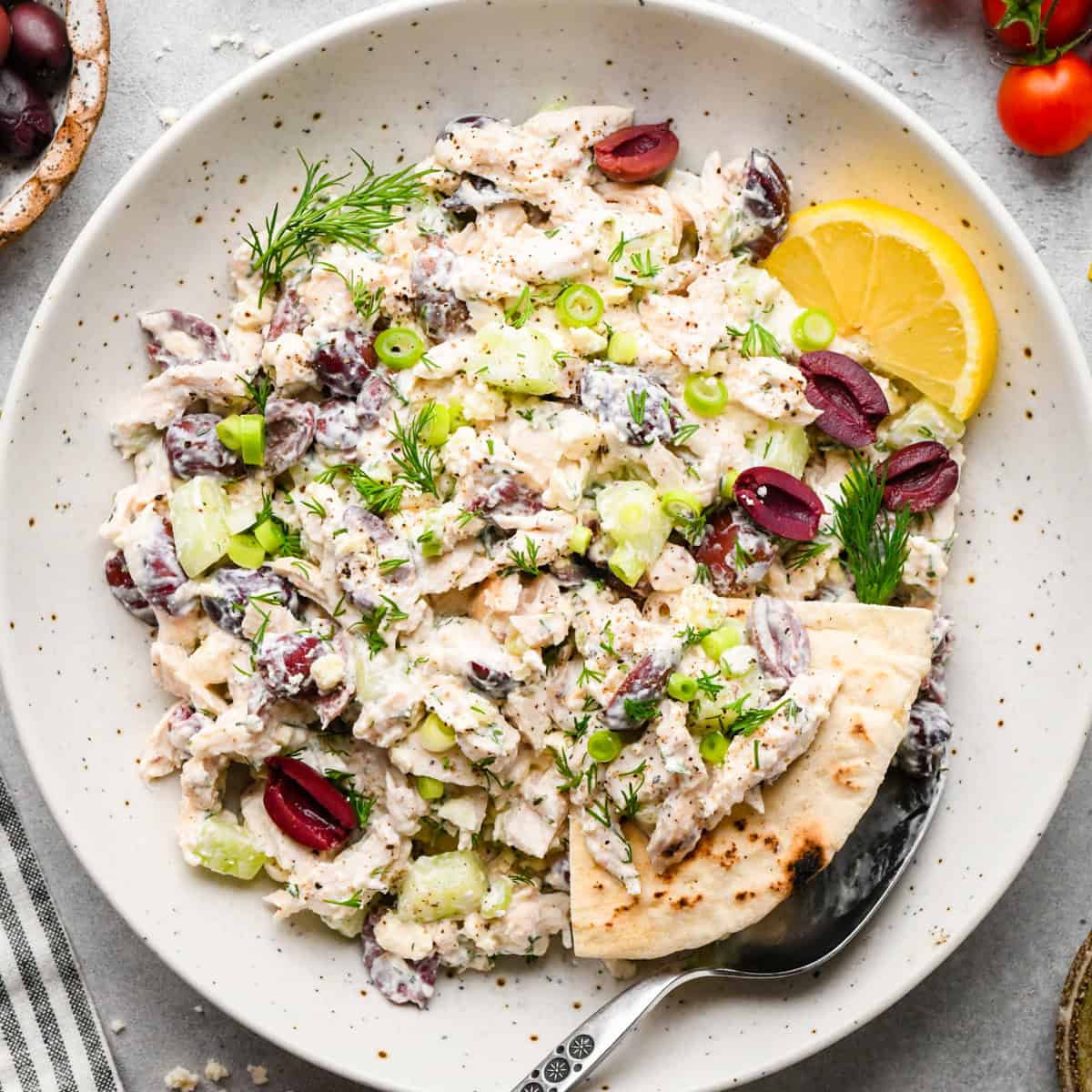 A bowl of Greek chicken salad made with shredded chicken, Kalamata olives, cucumber, green onions, and fresh dill mixed in a creamy dressing, served with pita bread and a lemon wedge on the side.