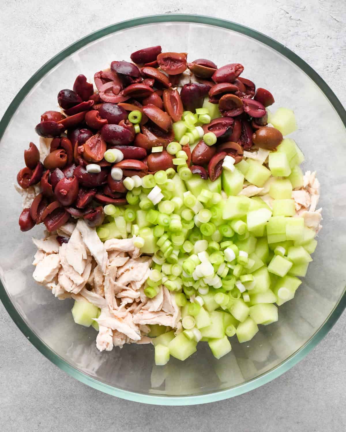 Making Greek chicken salad: kalamata olives, shredded chicken, cucumbers, and green onions are placed in a clear class bowl on a gray surface.