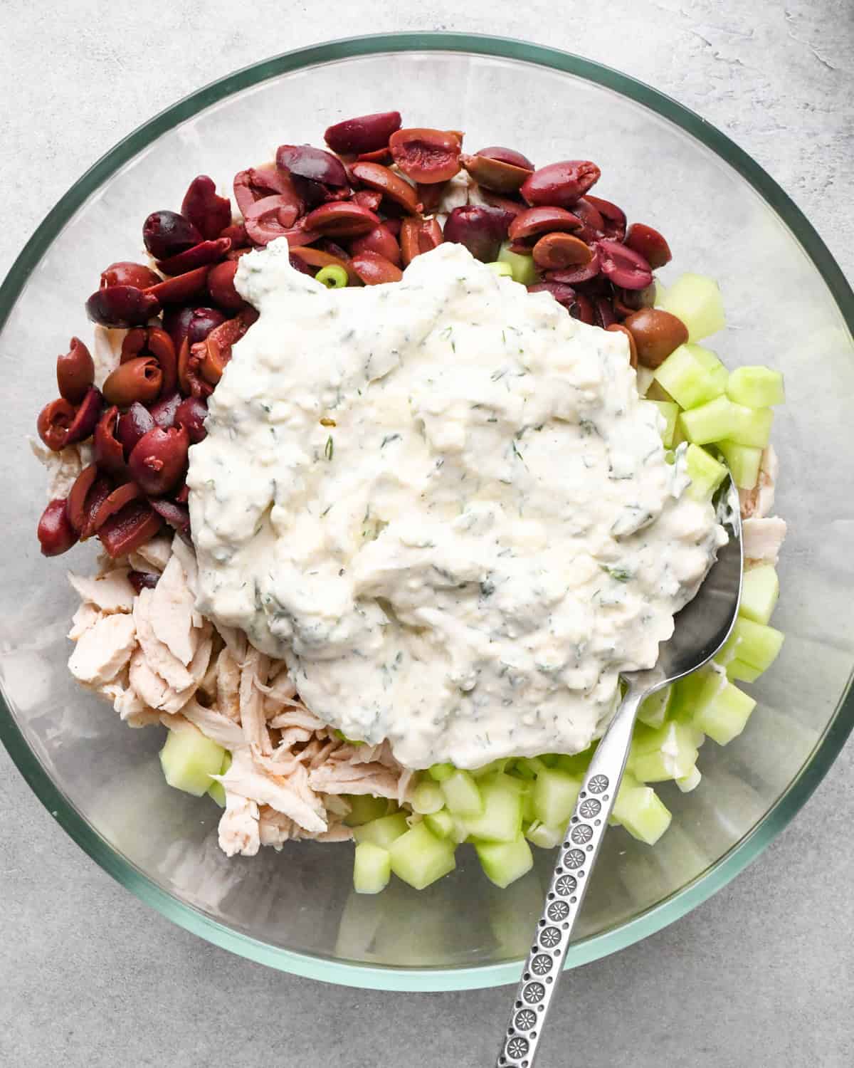 Making Greek chicken salad: kalamata olives, shredded chicken, cucumbers, green onions, and completed salad dressing are shown in a clear class bowl on a gray surface and being mixed together with a spoon.