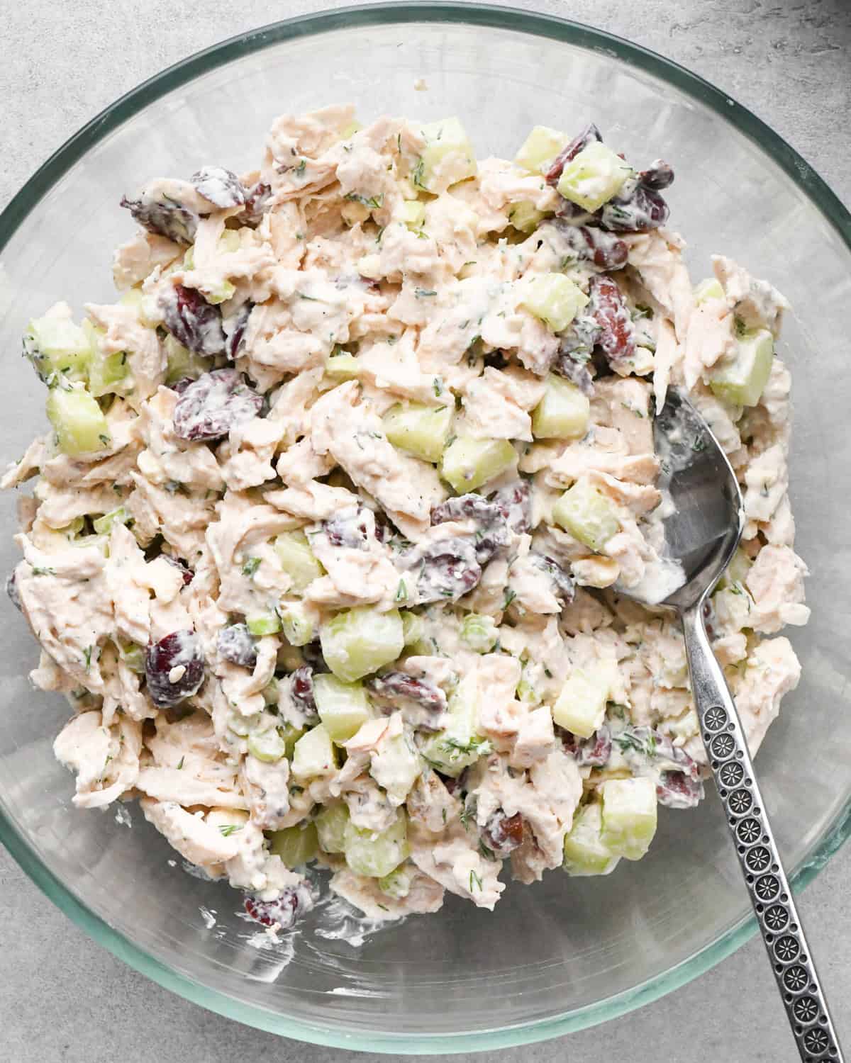 Making Greek chicken salad: kalamata olives, shredded chicken, cucumbers, and green onions are placed in a clear class bowl on a gray surface and mixed together.