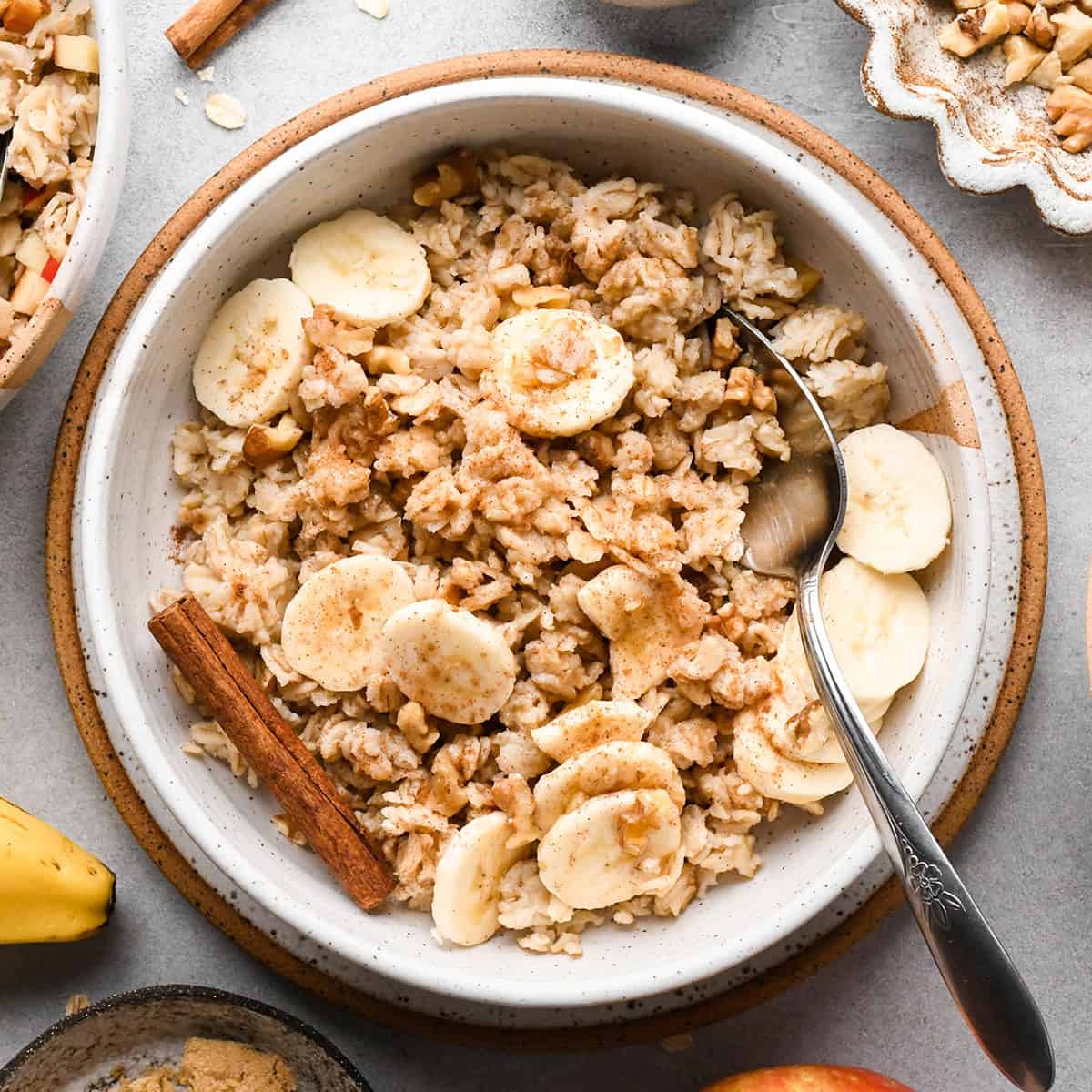 oatmeal in a bowl with bananas, cinnamon and a cinnamon stick. A hand is holding a silver spoon taking a btie.