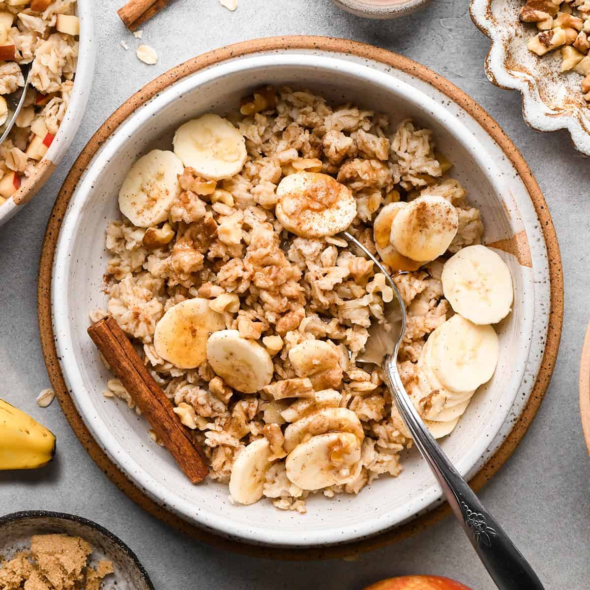 a bowl of oatmeal with sliced bananas and cinnamon. There's a spoon and a cinnamon stick in the oatmeal and it's surrounded by other bowls of oatmeal and ingredients.