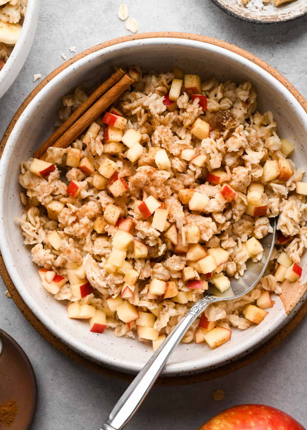 cooked oatmeal in a bowl with apples, cinnamon and a cinnamon stick. A hand is holding a silver spoon taking a bite.