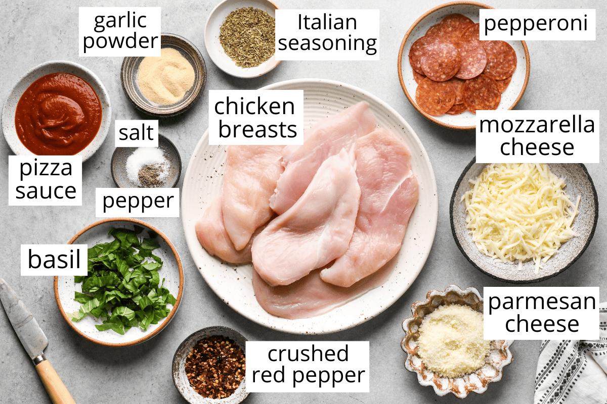 Top-down view of labeled ingredients for pizza chicken, including raw chicken breasts, pepperoni, mozzarella cheese, Parmesan cheese, pizza sauce, fresh basil, garlic powder, Italian seasoning, salt, pepper, and crushed red pepper in small bowls.