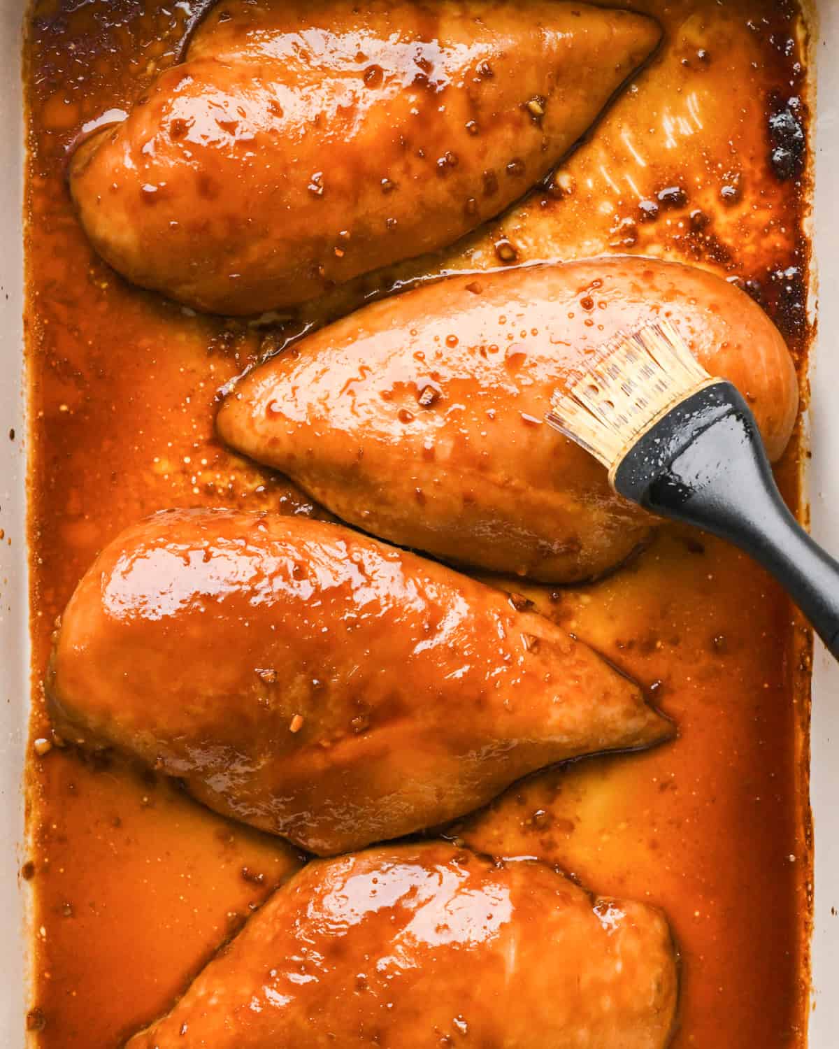 Making teriyaki chicken: marinated chicken breasts in a white casserole dish and are being brushed with extra sauce.