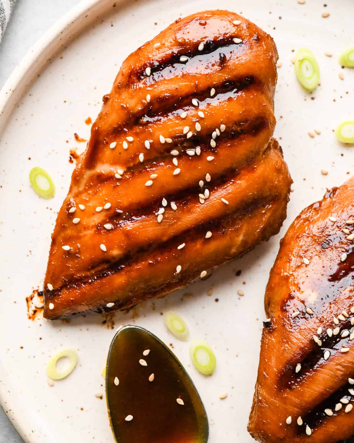 Grilled homemade teriyaki chicken breasts glazed with sauce, topped with sesame seeds and sliced green onions, served on a light-colored plate.