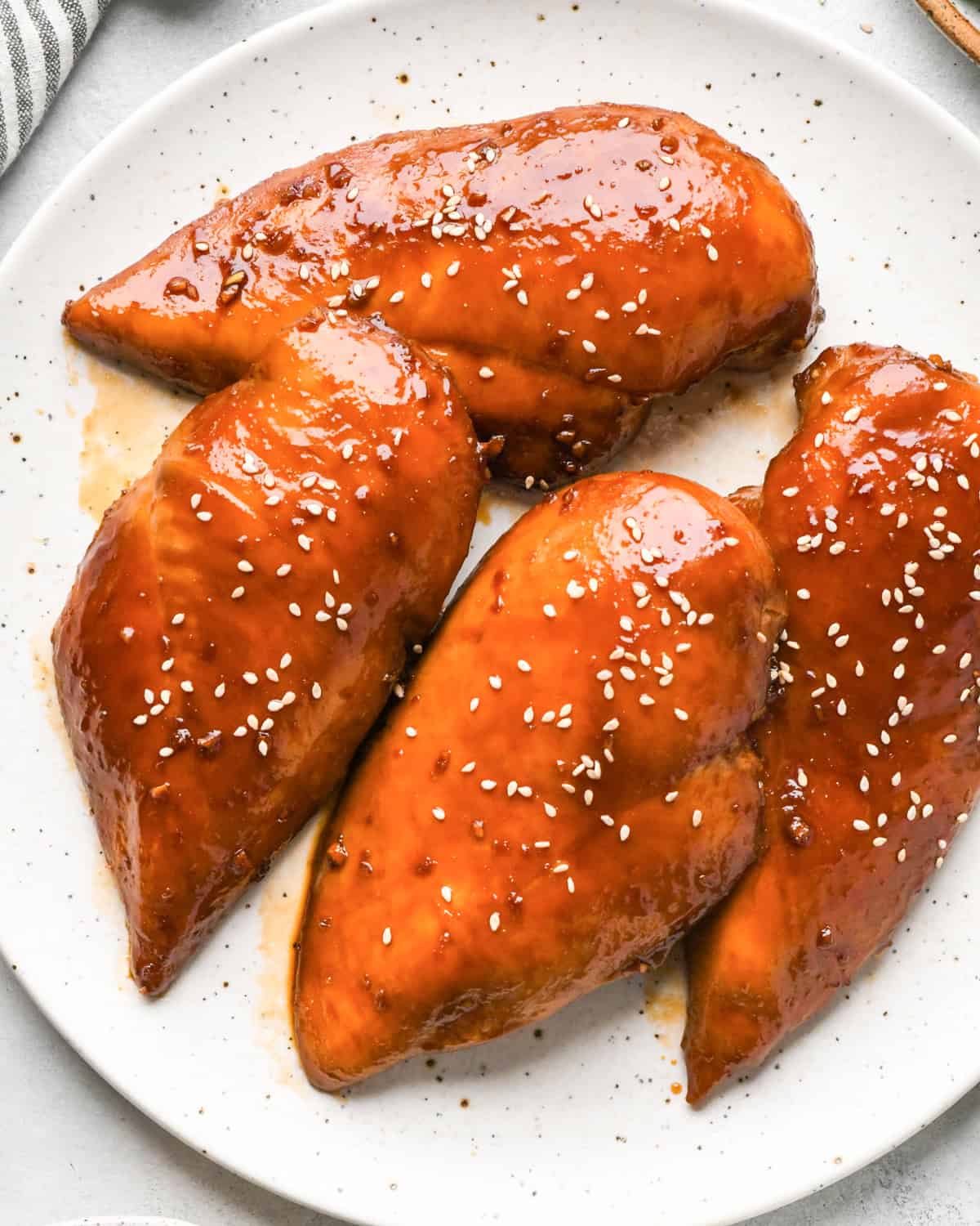 Grilled homemade teriyaki chicken breasts glazed with sauce, topped with sesame seeds and sliced green onions, served on a light-colored plate.