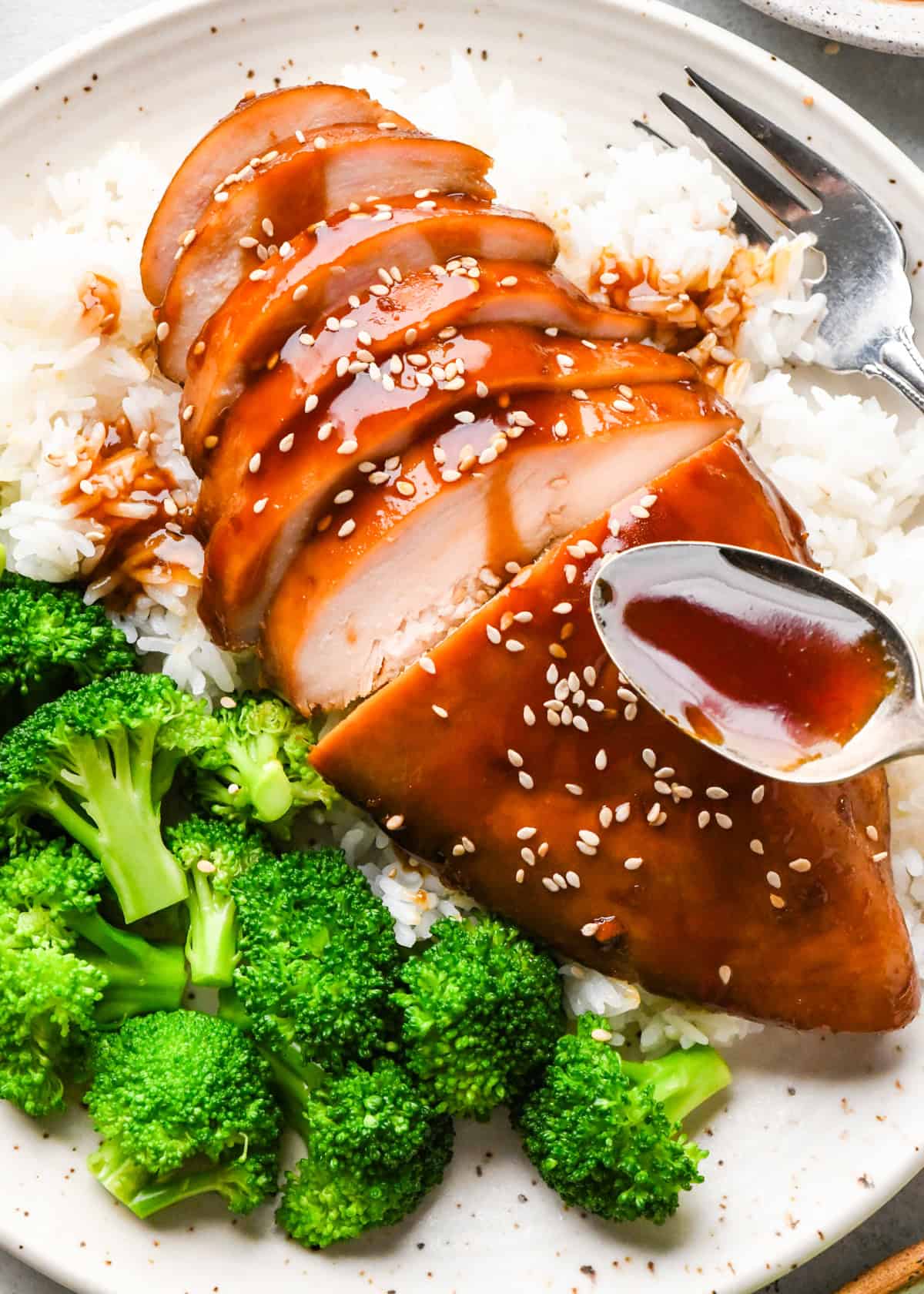 A plate of sliced homemade teriyaki chicken topped with sesame seeds and green onions, served over white rice with a side of steamed broccoli.