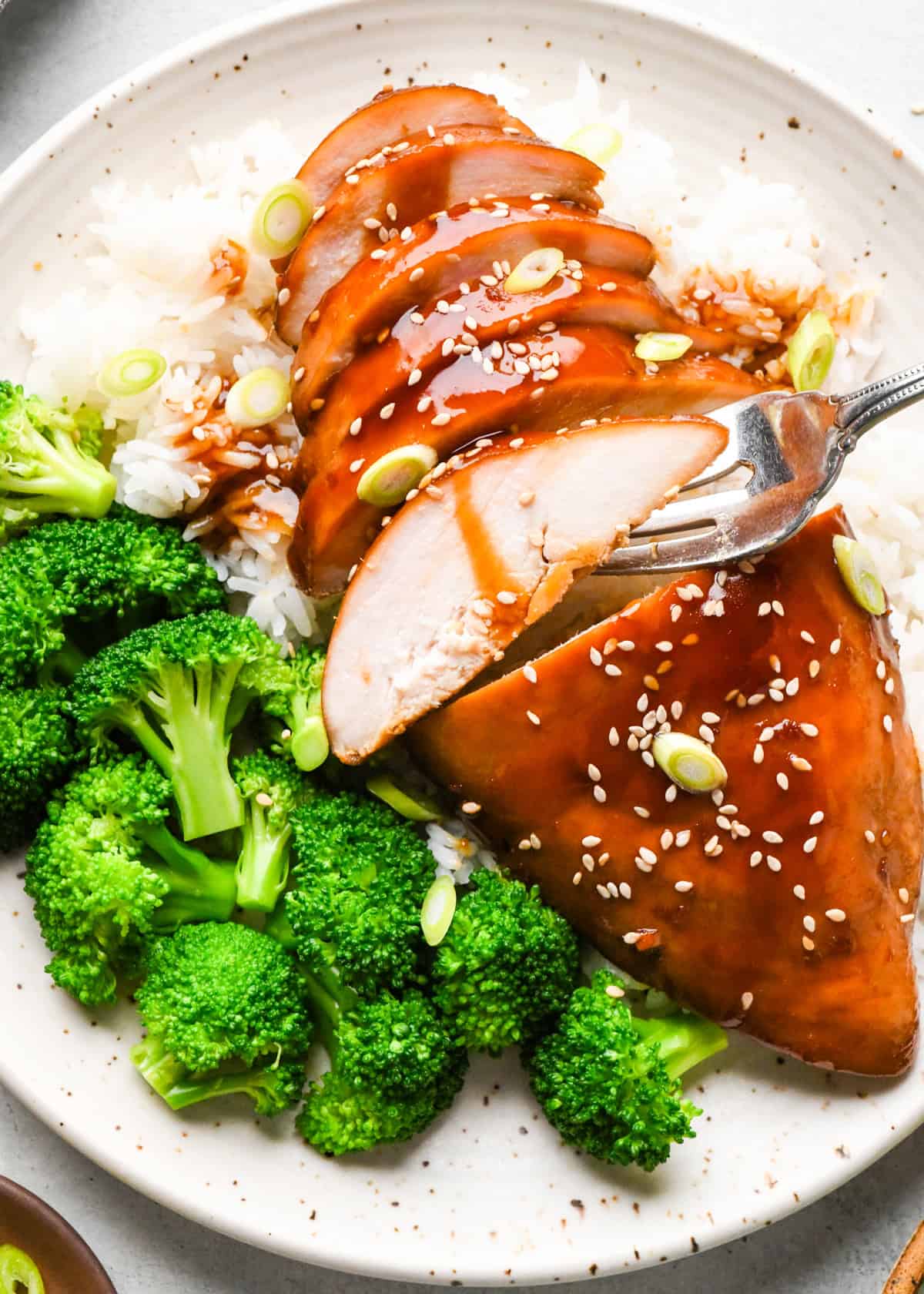 A plate of sliced homemade teriyaki chicken topped with sesame seeds and green onions, served over white rice with a side of steamed broccoli.