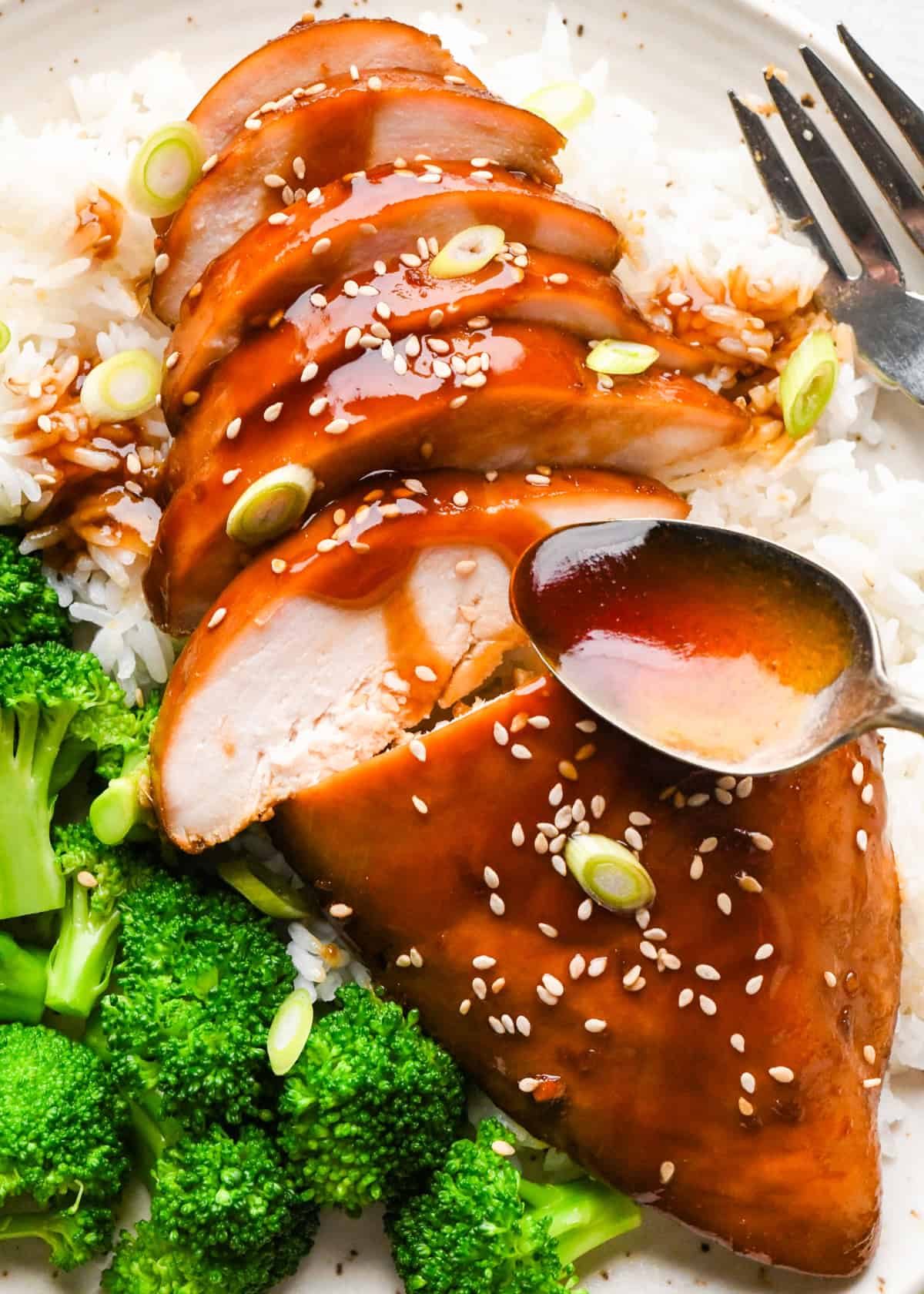 A plate of sliced homemade teriyaki chicken topped with sesame seeds and green onions, served over white rice with a side of steamed broccoli.