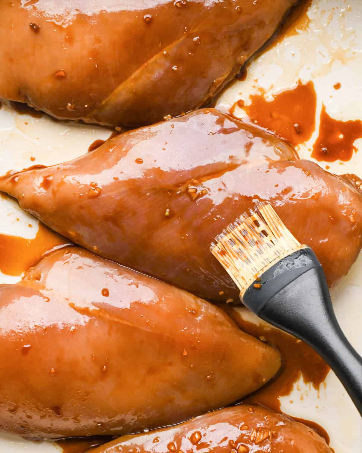 Making teriyaki chicken: marinated chicken breasts in a white casserole dish and are being brushed with extra sauce.
