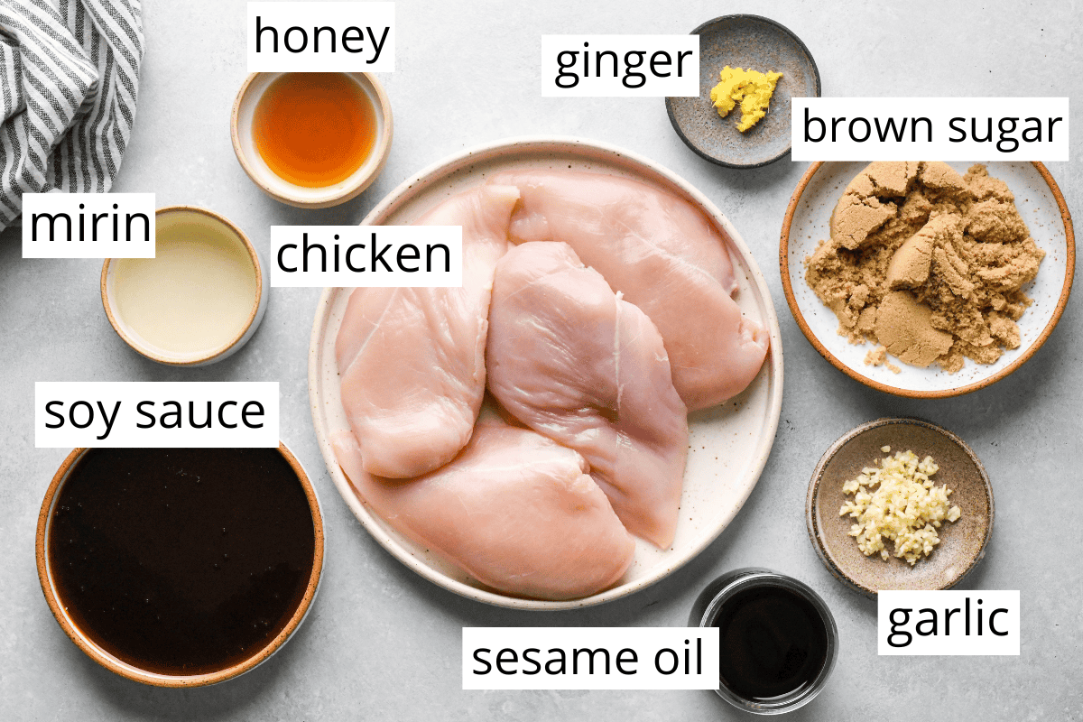 Top-down view of labeled ingredients for homemade teriyaki chicken, including raw chicken breasts, soy sauce, mirin, honey, brown sugar, sesame oil, garlic, and ginger, arranged neatly on a light countertop.