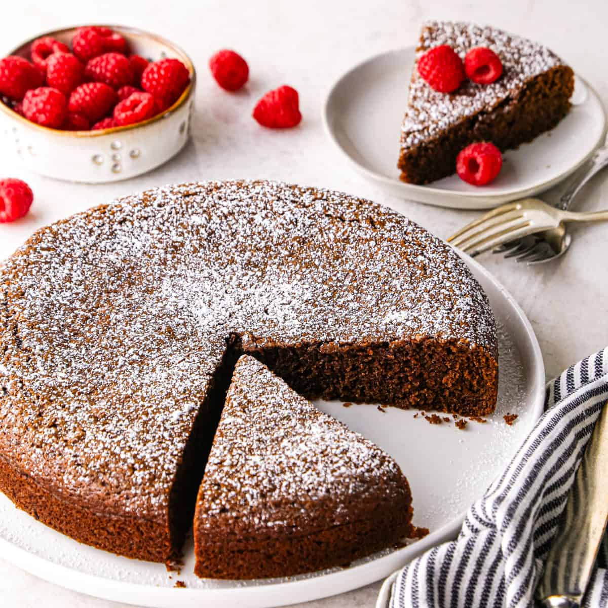 a round chocolate olive oil cake topped with powdered sugar on a white cake plate. Two pieces are cut out of it, one is still on the cake plate and one is on a small round white plate in the background topped with raspberries. There is a bowl of raspberries in the top left corner and a striped towel in the bottom right corner.