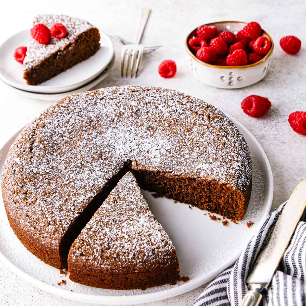 a round chocolate olive oil cake topped with powdered sugar on a white cake plate. Two pieces are cut out of it, one is still on the cake plate and one is on a small round white plate in the background topped with raspberries. There is a bowl of raspberries in the top right corner and a striped towel in the bottom right corner.