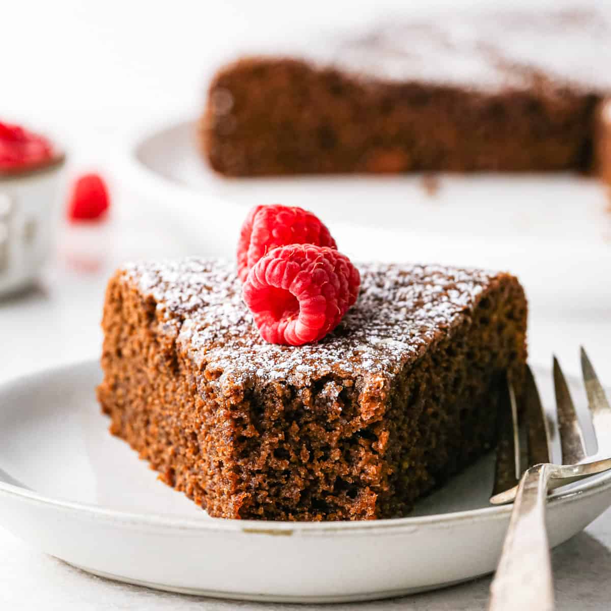a piece of chocolate and olive oil cake on a plate with a bite taken out of it and a fork on the right side of the cake on the plate. It's topped with powdered sugar and two raspberries. The rest of the cake and a bowl of raspberries are in the background