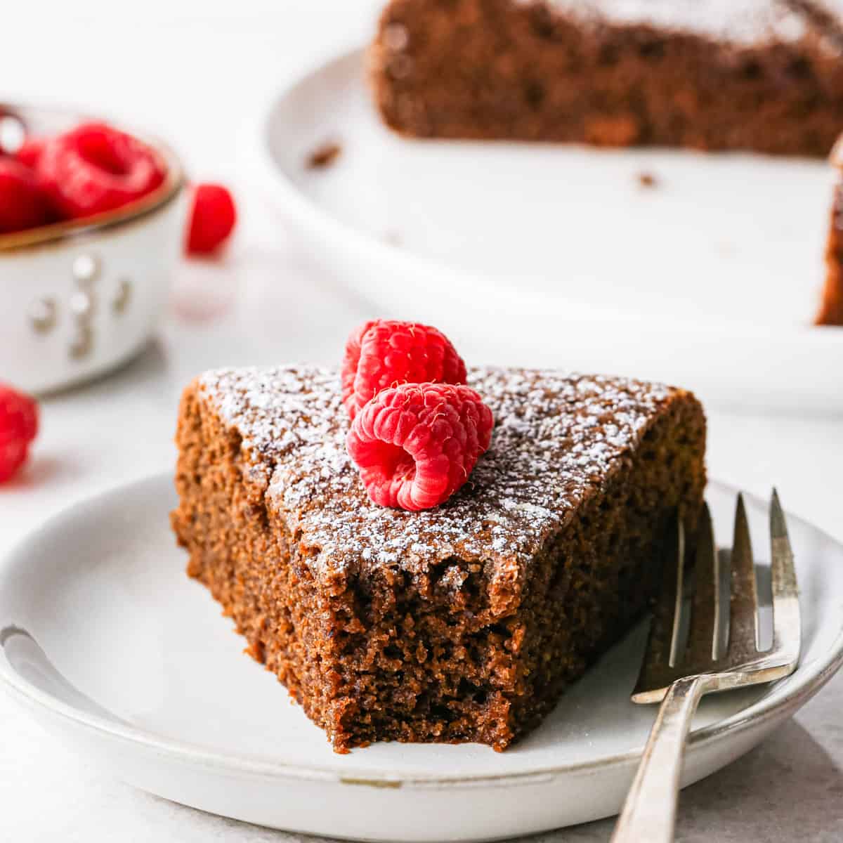 a piece of chocolate and olive oil cake on a plate with a bite taken out of it and a fork on the right side of the cake on the plate. It's topped with powdered sugar and two raspberries. The rest of the cake and a bowl of raspberries are in the background