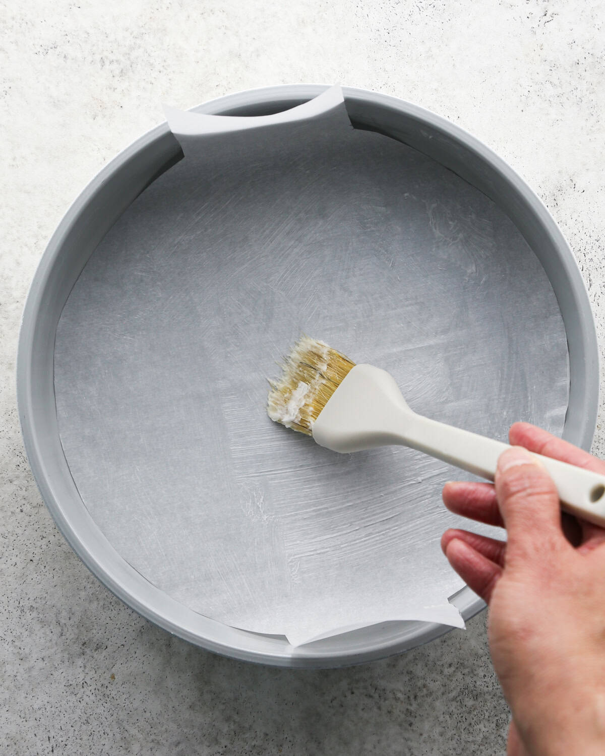butter being brushed over parchment paper in a round silver cake plate