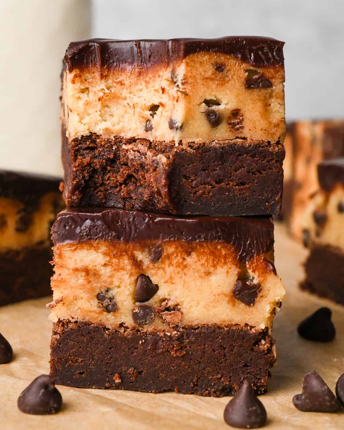 stack of two cookie dough brownies with a fudgy chocolate brownie on the bottom, edible chocolate chip cookie dough in the middle, and a glossy chocolate ganache on top on parchment paper. The top brownie has a bite taken out fo it.  Surrounded by other cookie dough brownies and chocolate chips